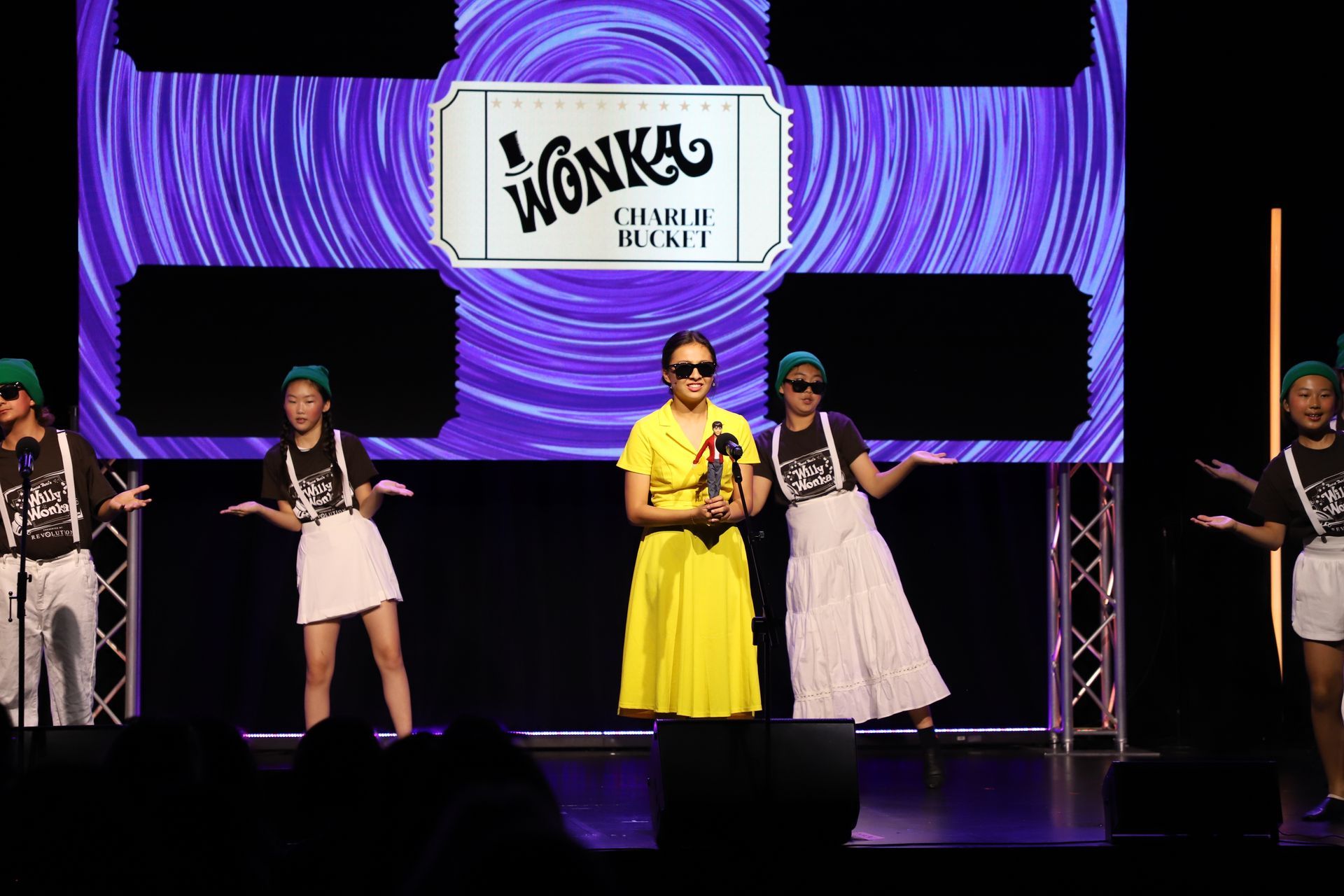 Four people pose on stage in colorful outfits beneath a purple Wonka-themed backdrop for a theater performance.