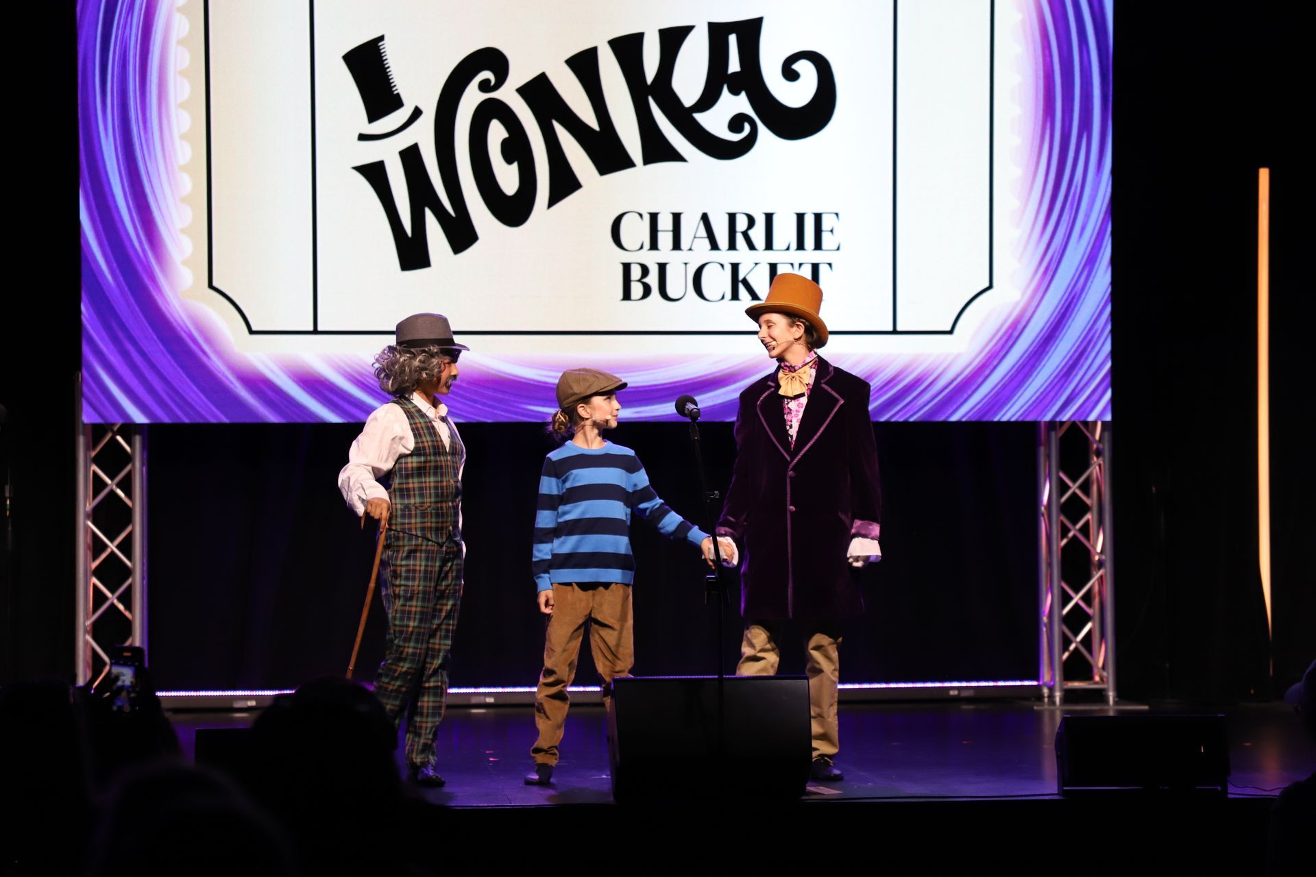 Three performers on stage under a Charlie and the Chocolate Factory sign, dressed in costumes.