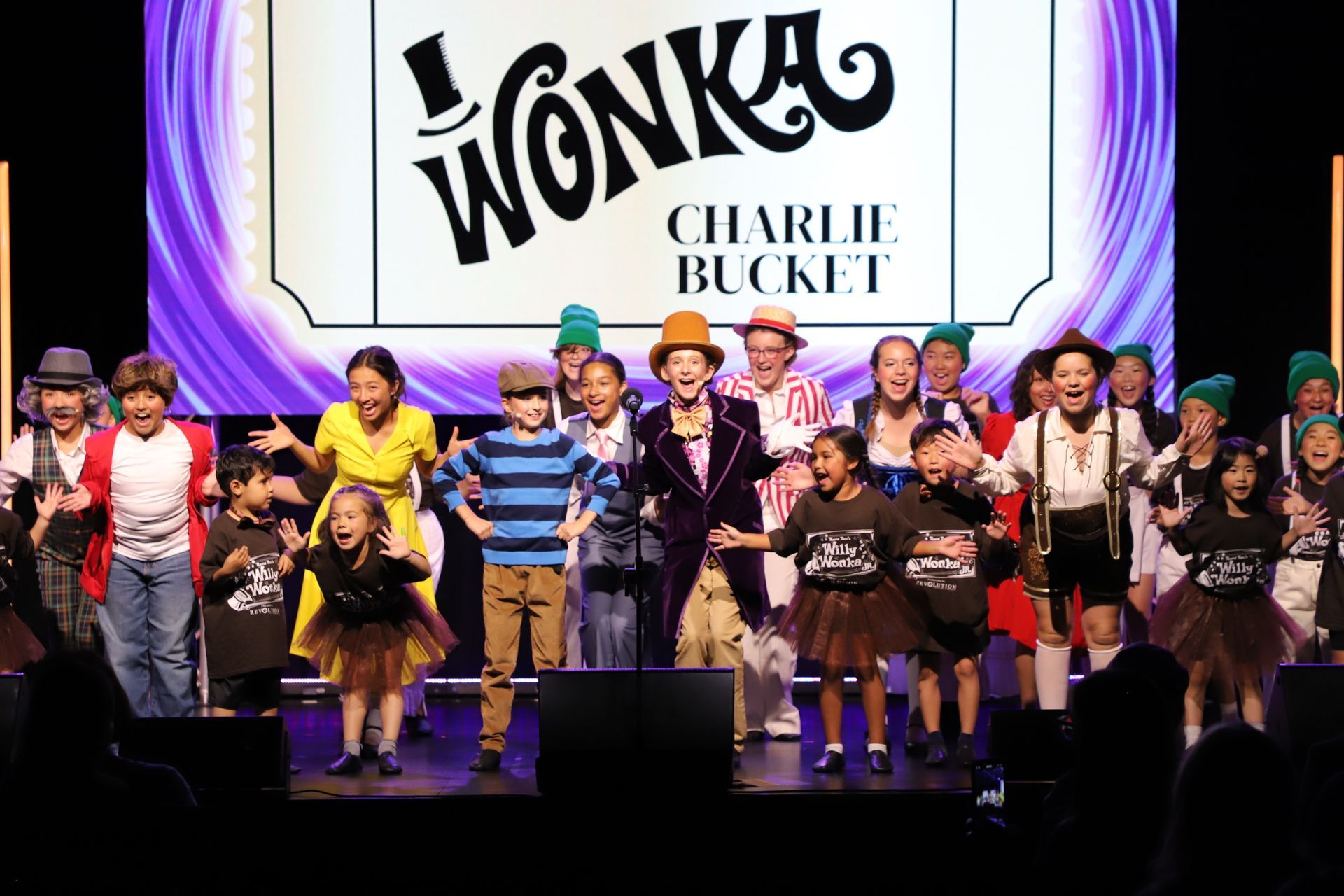 Children in costume on a stage under a large “Wonka” sign, performing in a theatrical group scene