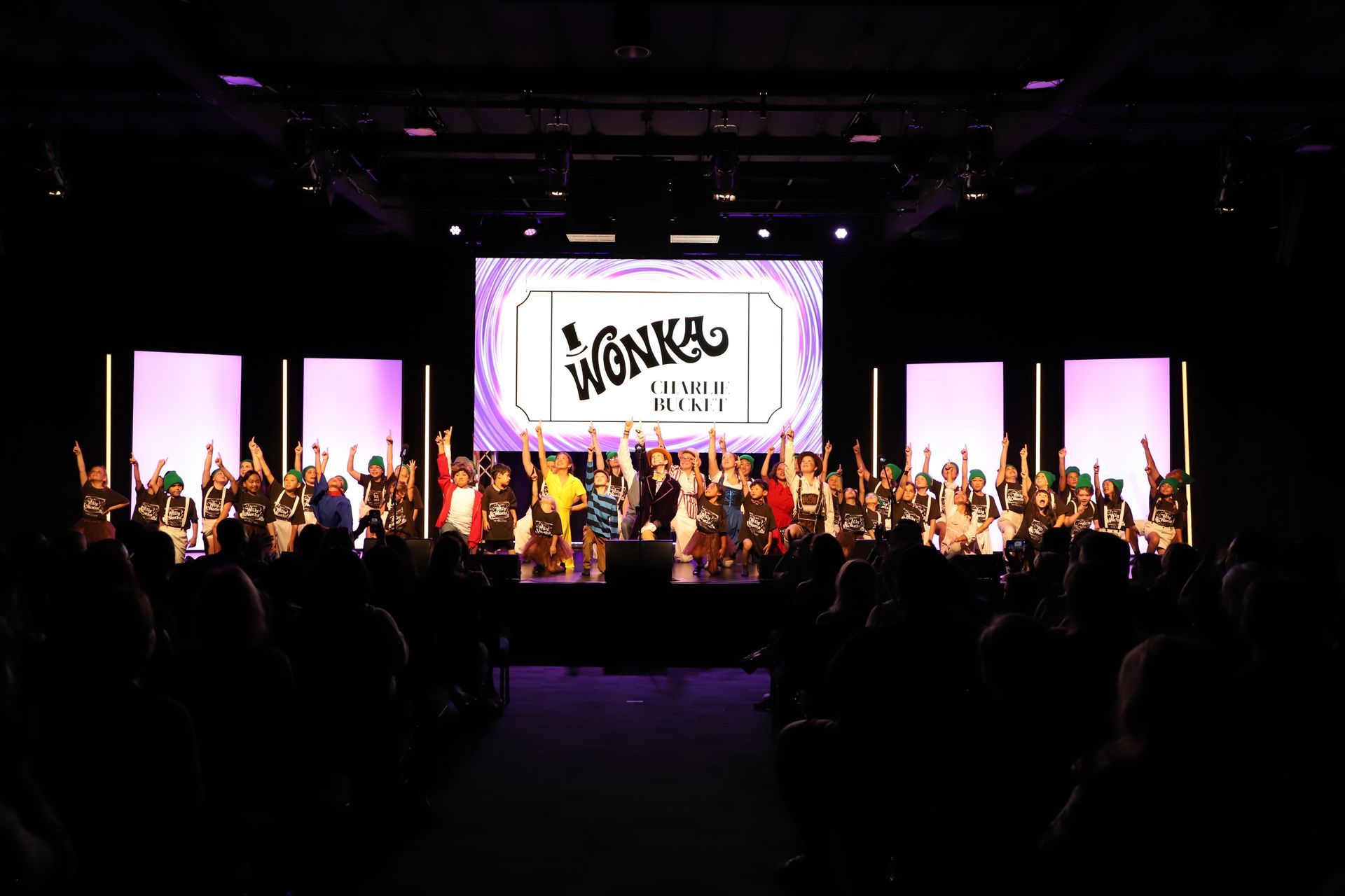 Large choir performing on a lit stage with a “SANCTUARY” sign, seen from a dark audience hall for a theater performance.