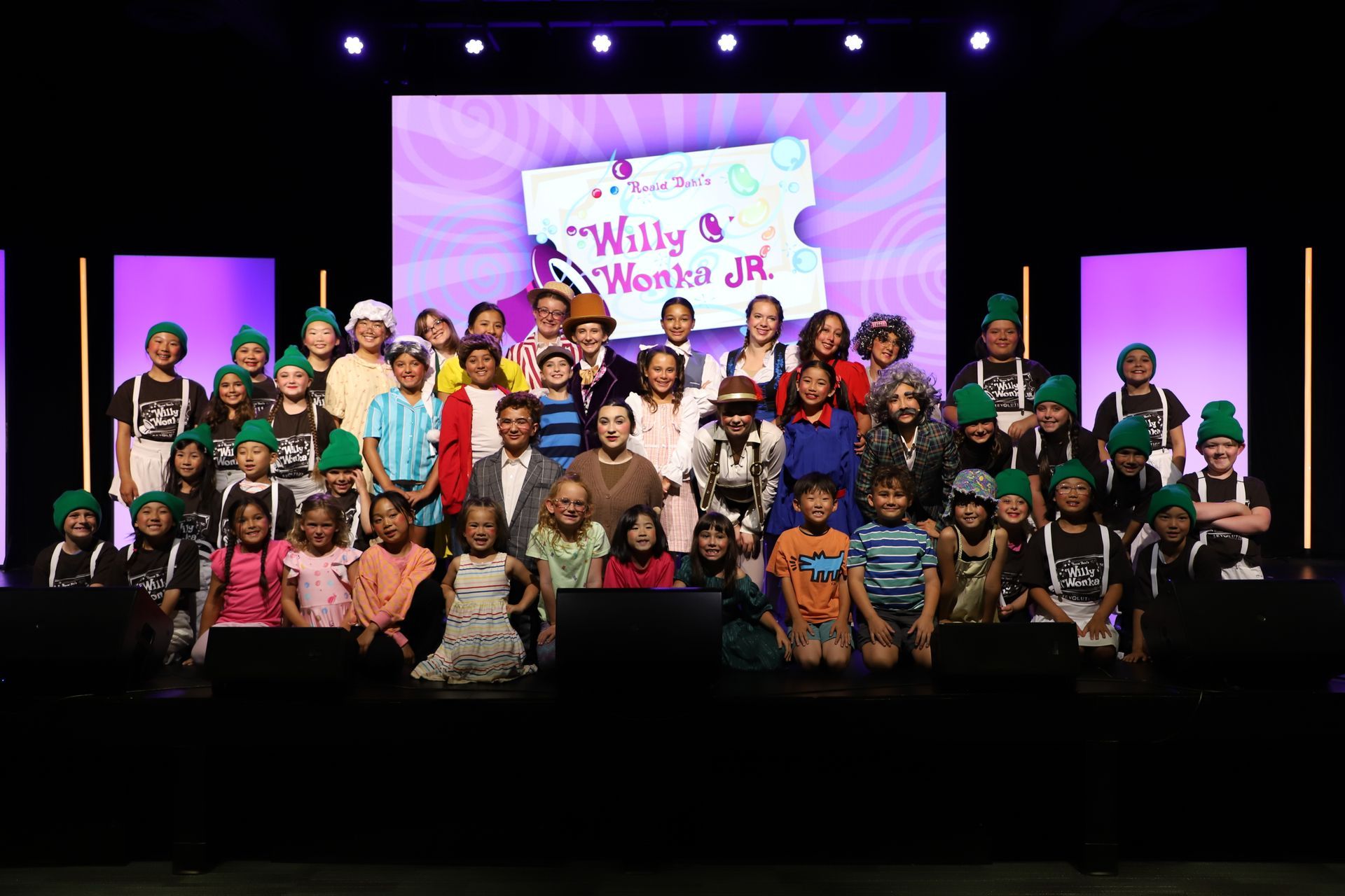Large group posing on a stage in front of a colorful screen that reads “Willy & Wen Jr” for a theater performance.