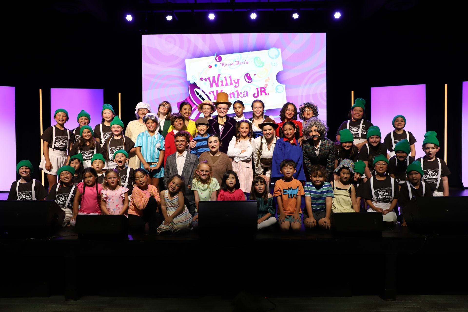 Large group posing on a stage with purple lights and a decorated screen backdrop for a theater performance.