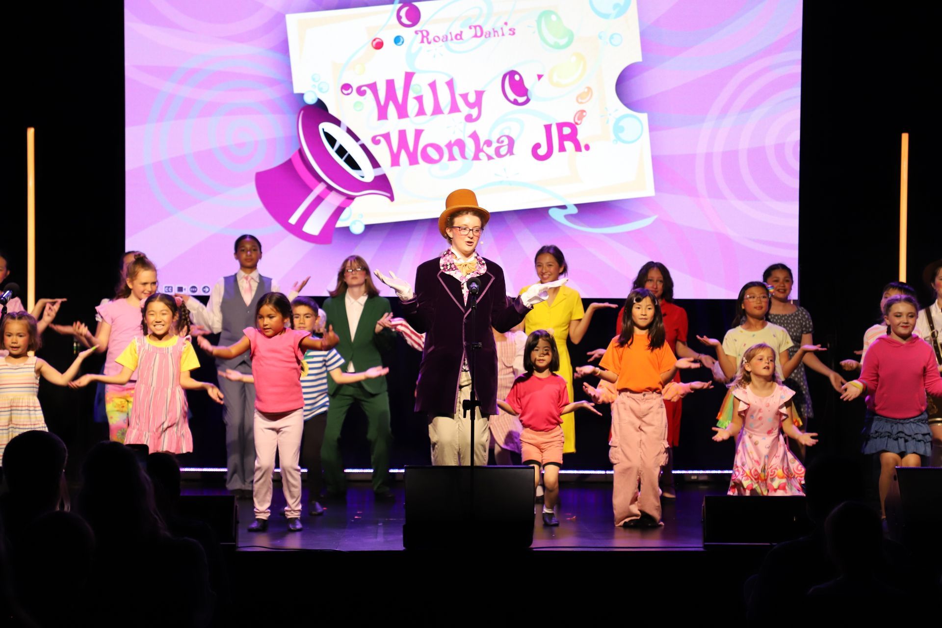 Children performing on stage with a host in front of a Willy Wonka Jr. backdrop for a theater performance.