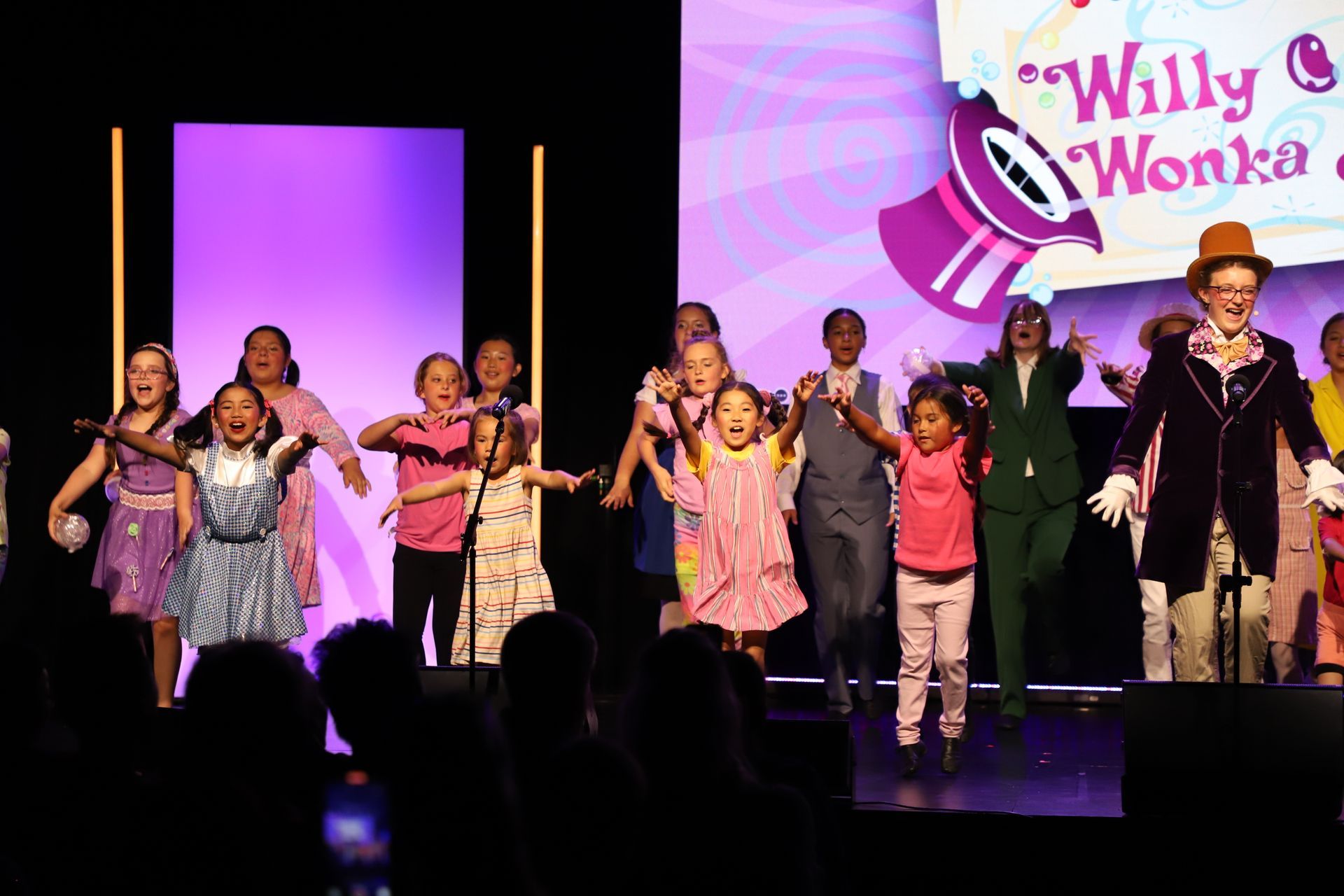 Children performing on a stage with purple lights, wearing colorful costumes in a Willy Wonka-themed show.