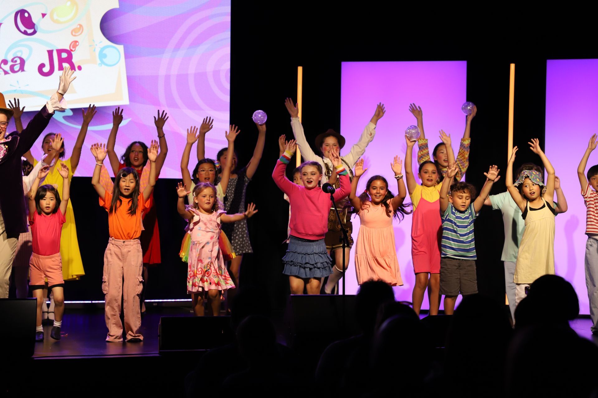 Children performing on a lit stage, raising their arms during a lively dance recital for a theater showcase.