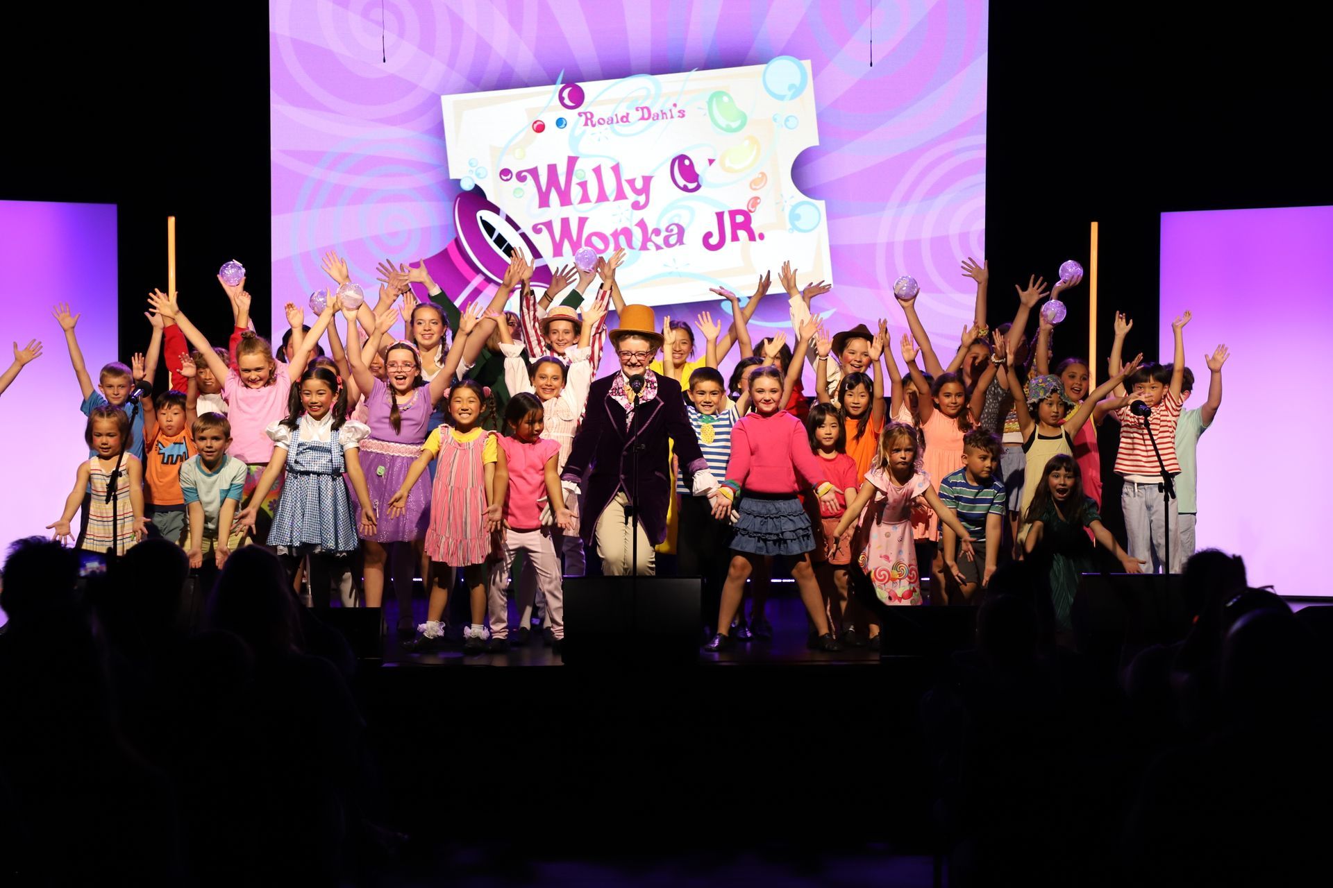 Children on stage with raised arms under pink lights, in front of a 