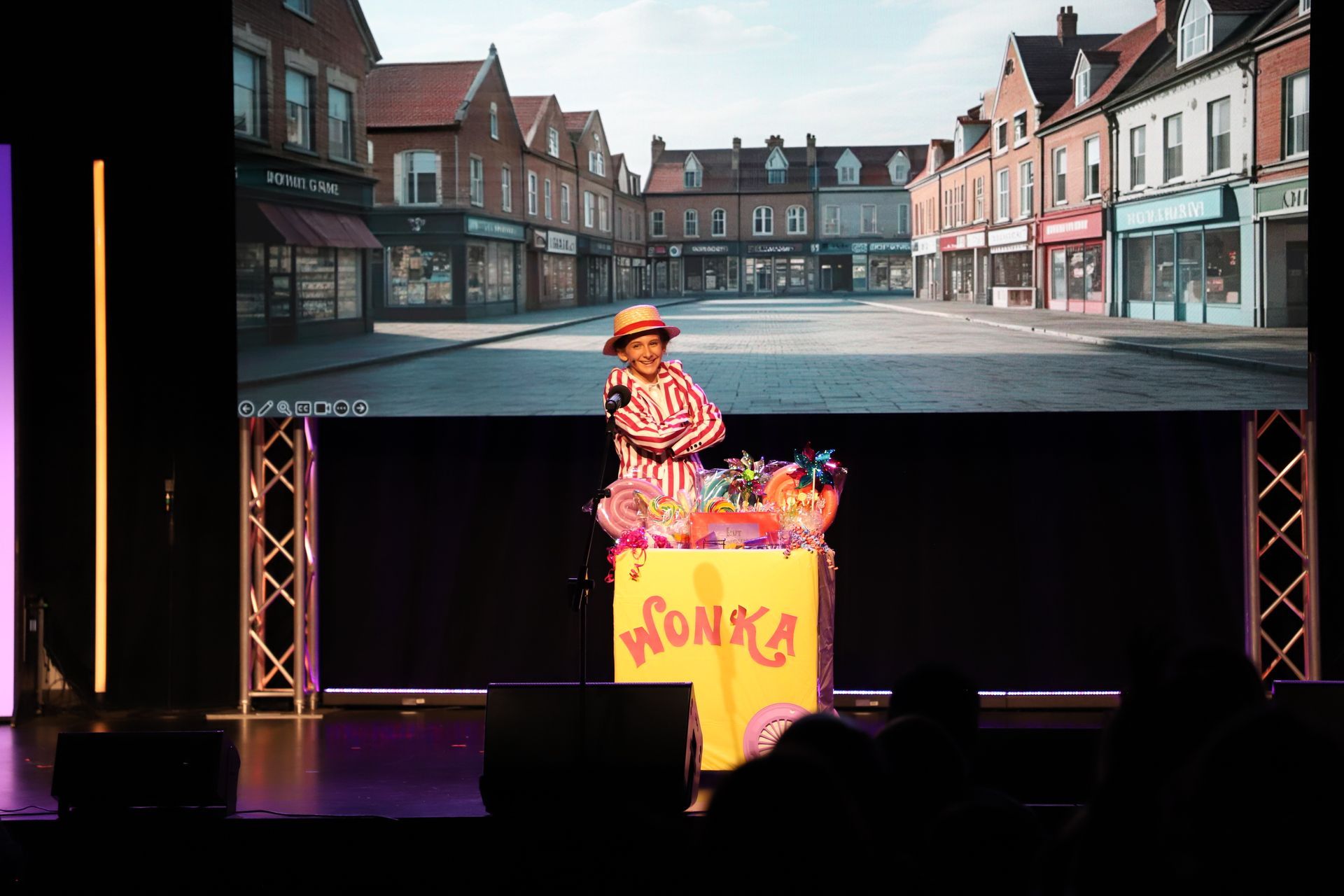Person on stage behind a pink podium, with a street-scene backdrop and audience silhouettes in front for a theater showcase.
