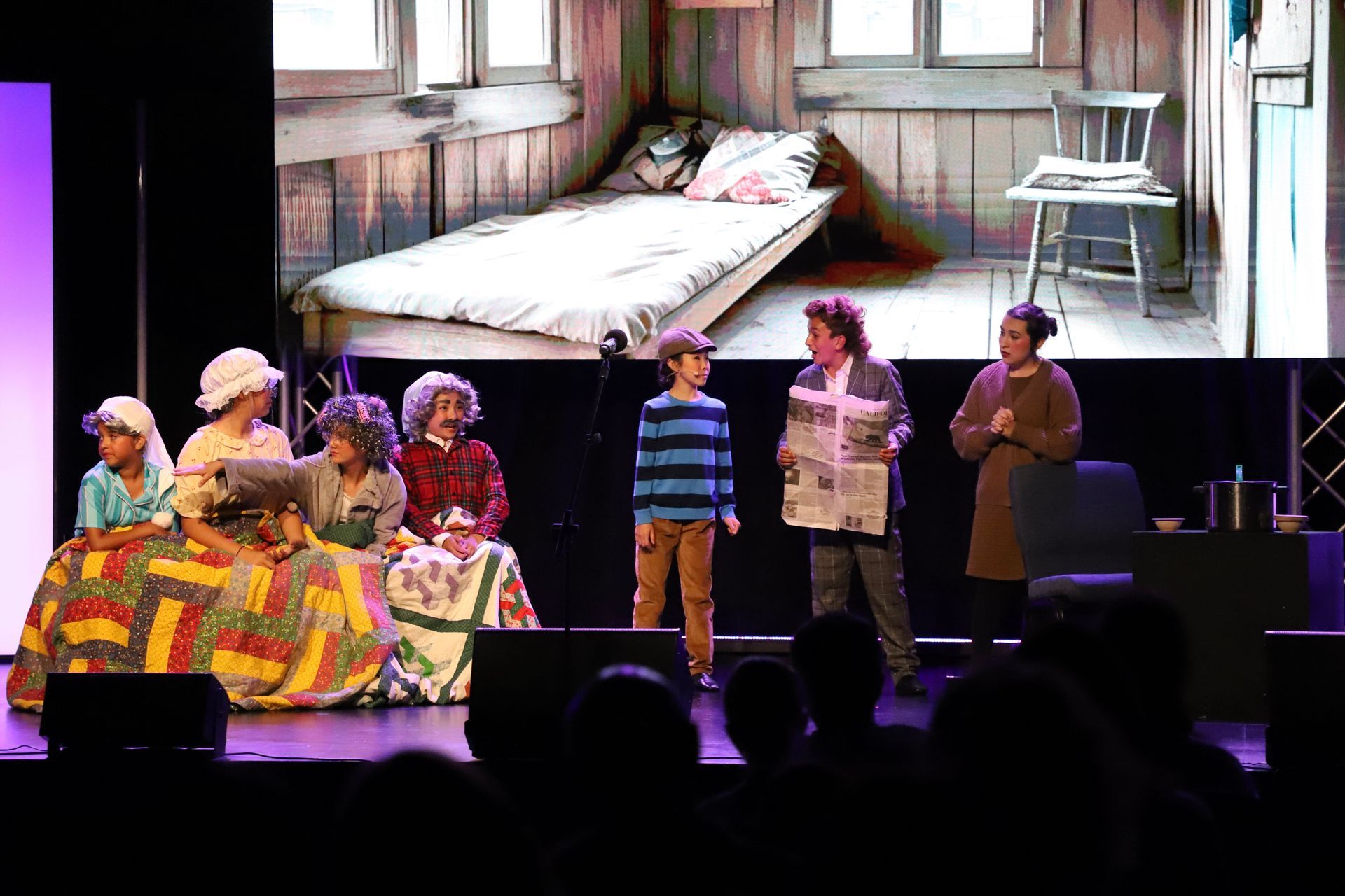 People on a stage in front of a bedroom set backdrop, with audience silhouettes in the foreground for a theater showcase.