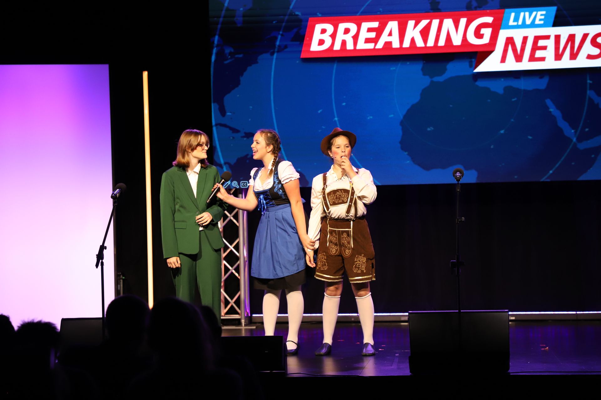 Three people on stage under a “Breaking News” screen, one speaking into a microphone for a theater showcase.