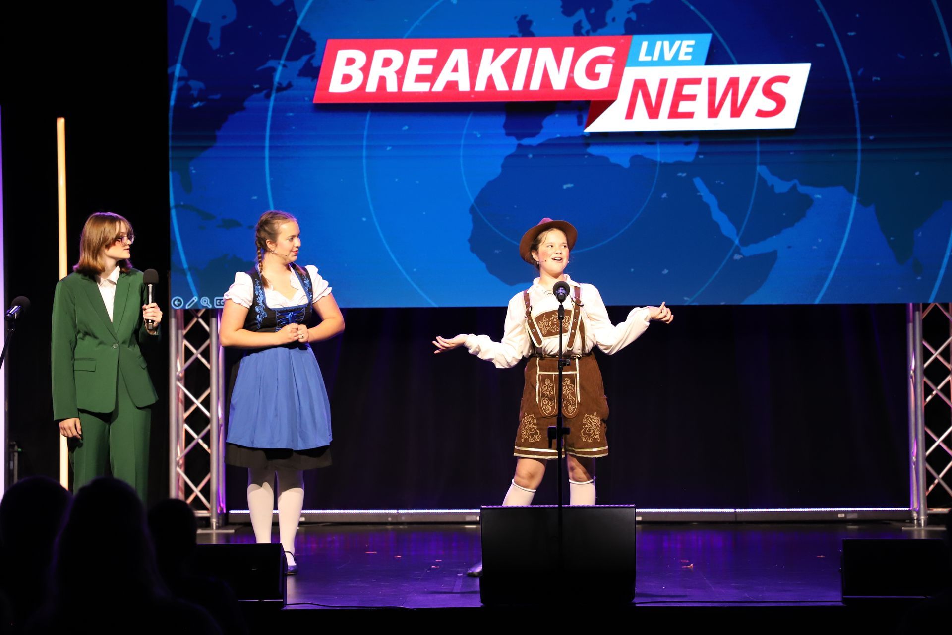 Three performers onstage under a “Breaking News” screen, with microphones and bright stage lighting for a theater showcase.