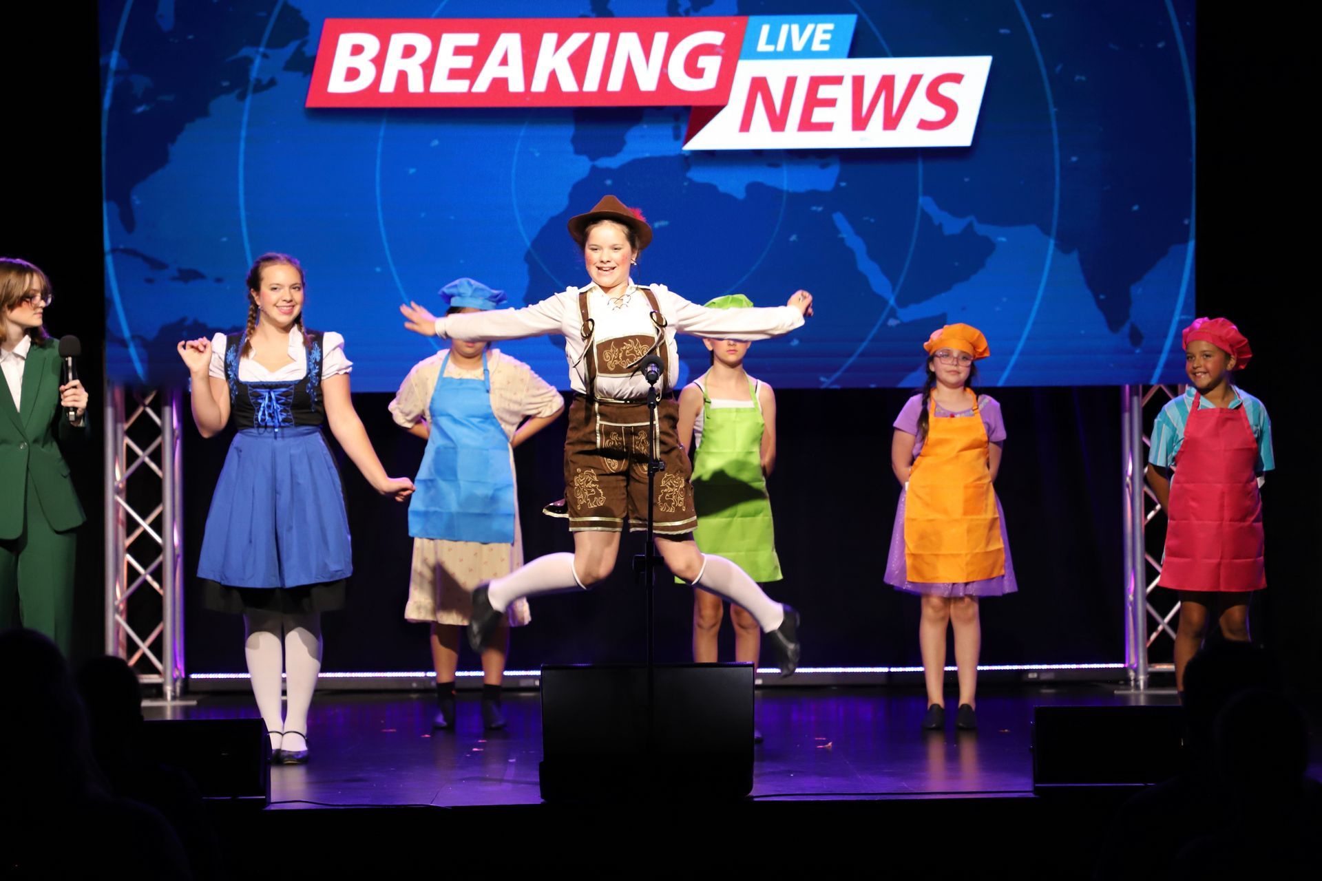 Performers on stage under a “Breaking Live News” sign  posing in a theatrical scene for a theater performance.