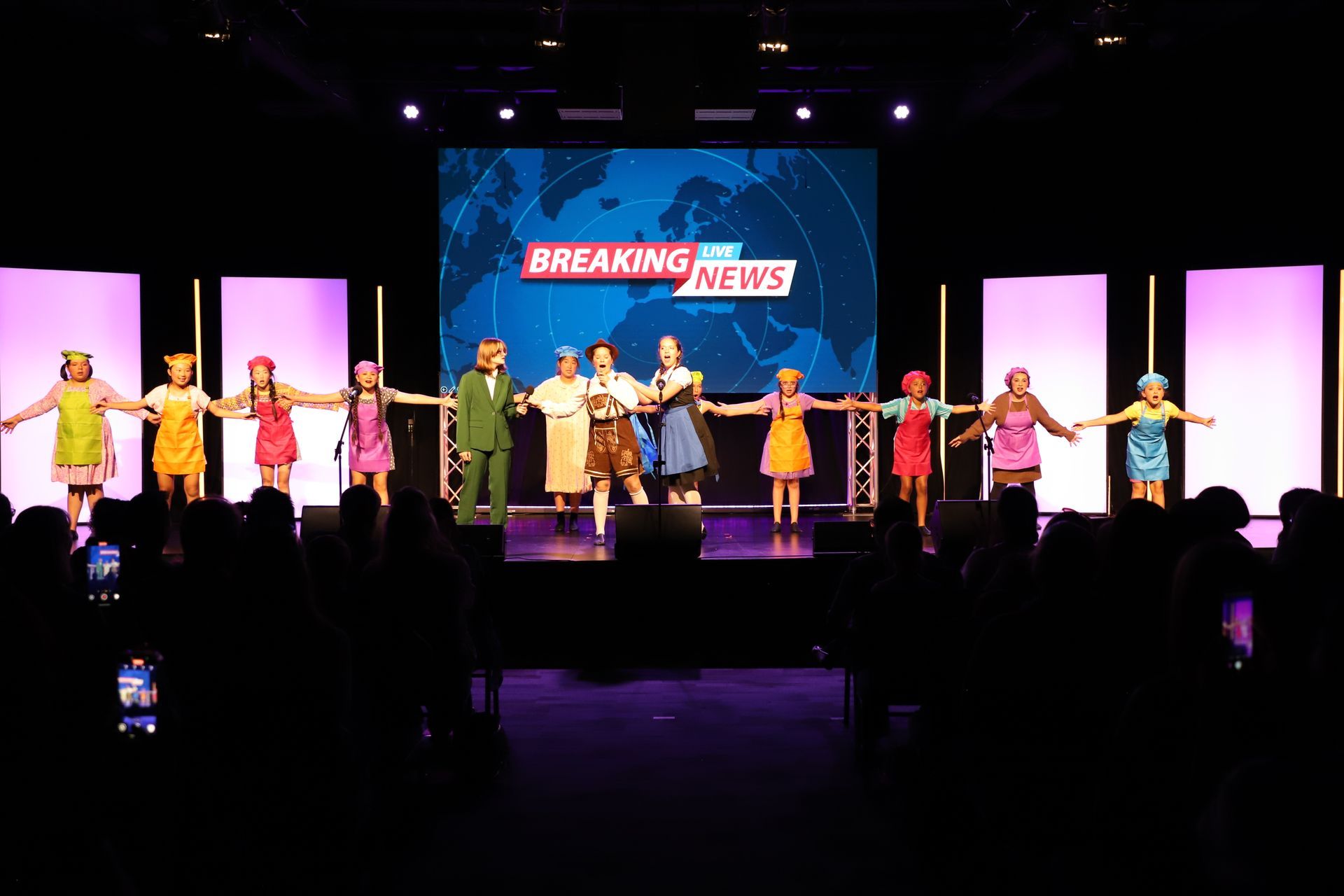 Stage performance with dancers in colorful costumes beneath a “BREAKING NEWS” backdrop for a theater performance.