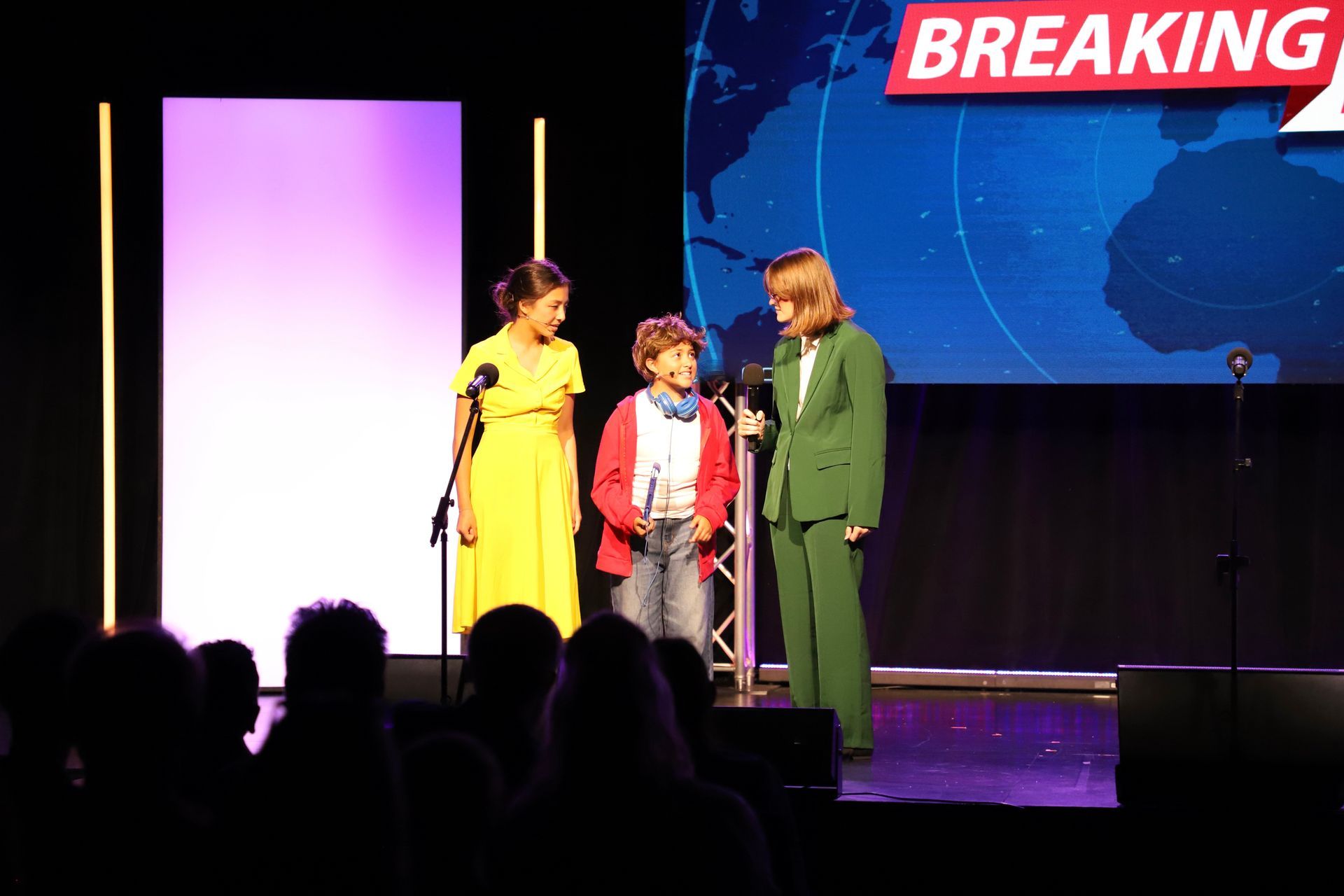 Three people on stage speaking into microphones, with a “BREAKING” screen in the background for a theater performance.