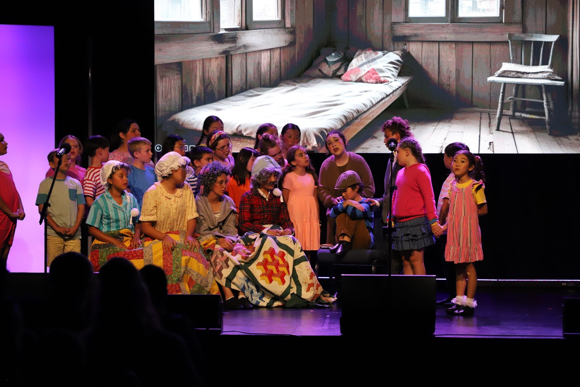 Children in colorful costumes perform onstage before a projected bedroom backdrop for a theater performance.