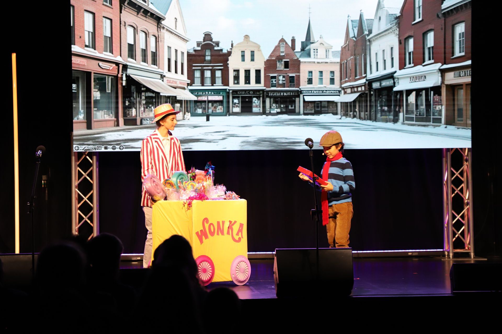 Two people in costumes on a stage, with a snowy village scene projected behind them for a theater performance.