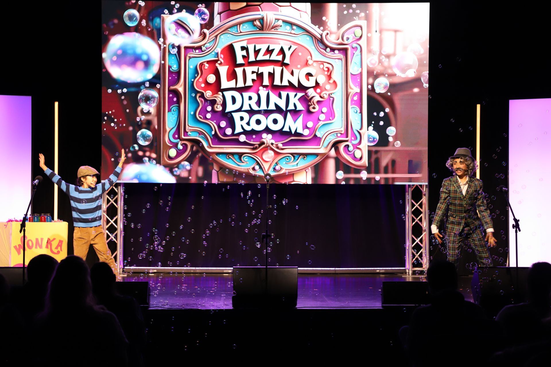 Two performers on a lit stage with a neon “Drinking Room” sign for a theater performance.
