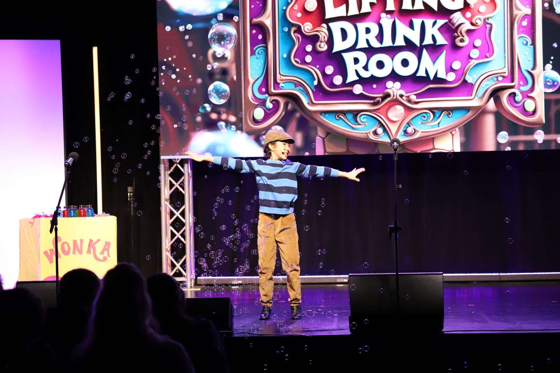 Performer on stage with arms outstretched beneath a colorful “Drink Room” sign for a theater performance.