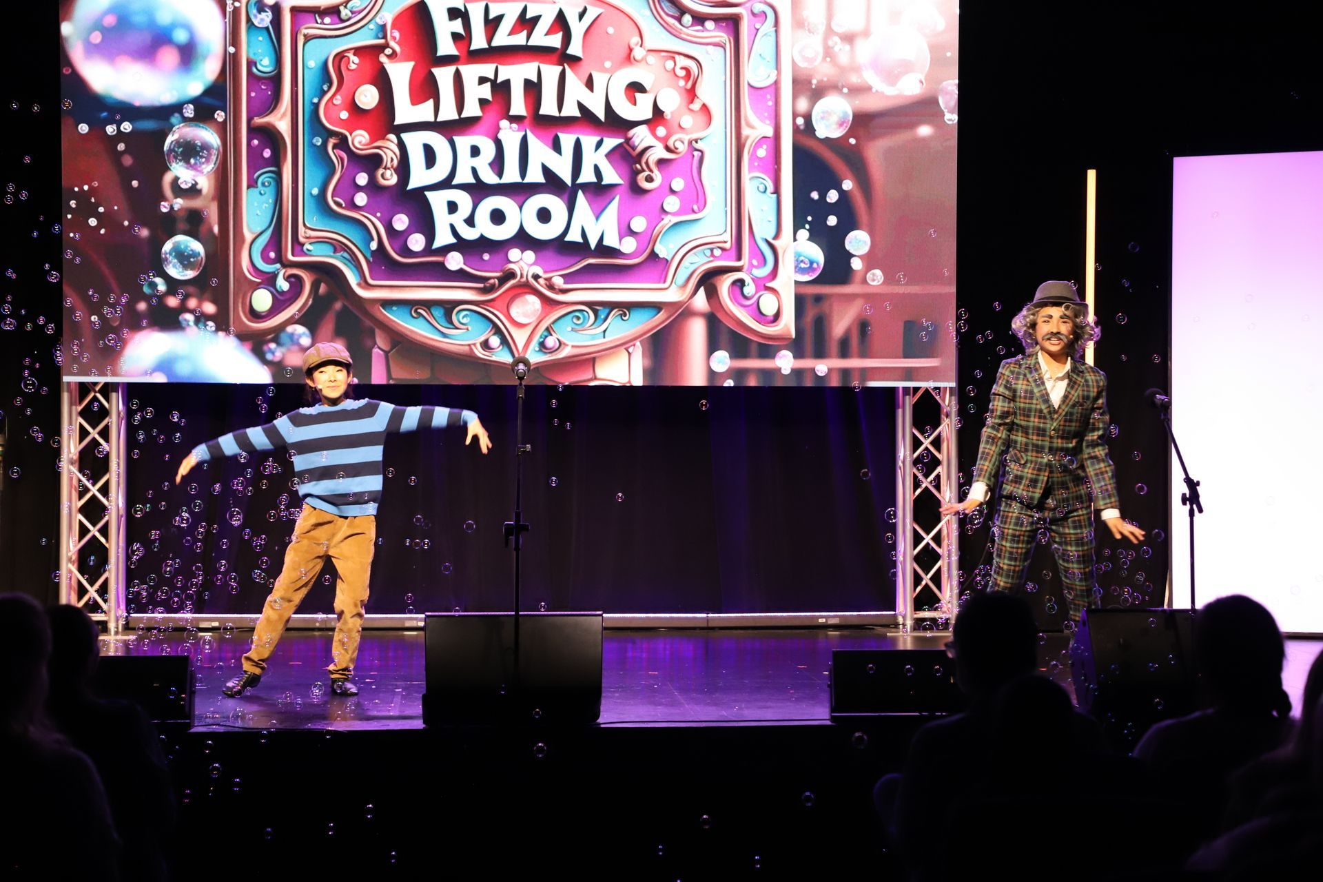Two performers onstage under a “Fizzy Lifting Drink Room” sign for a theater performance.