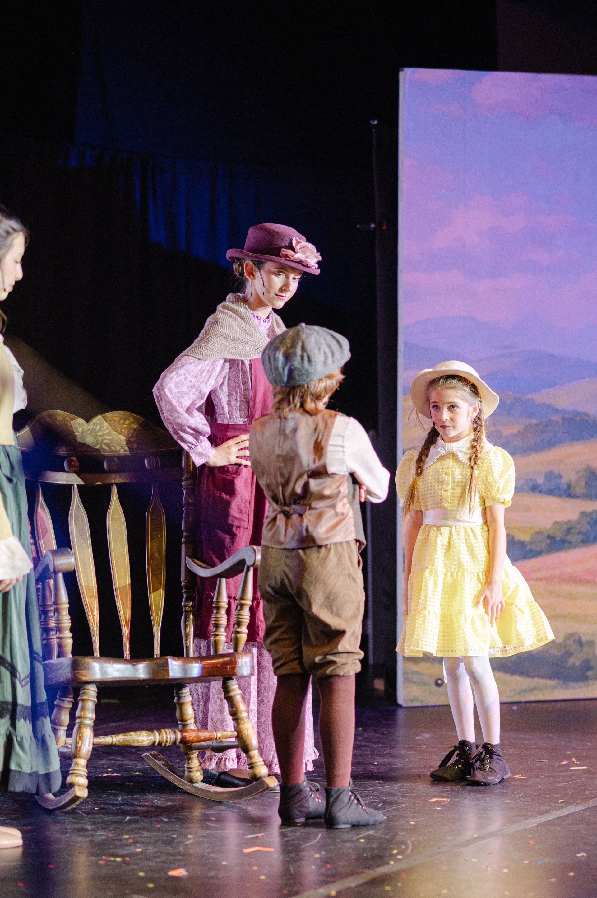 Children on a stage in costume, with a painted backdrop and props from The Music Man
