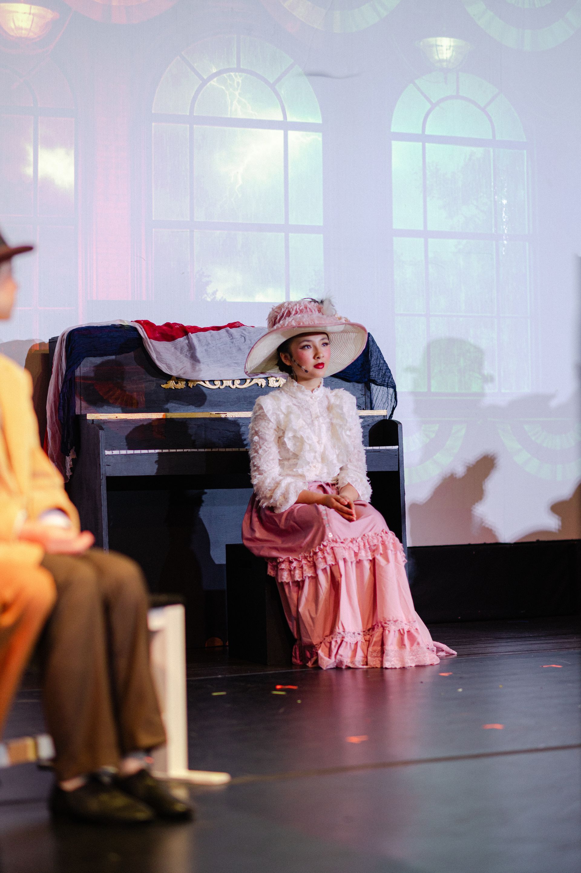 Child in a hat and pink dress seated onstage beside a piano, with a projected window backdrop from The Music Man