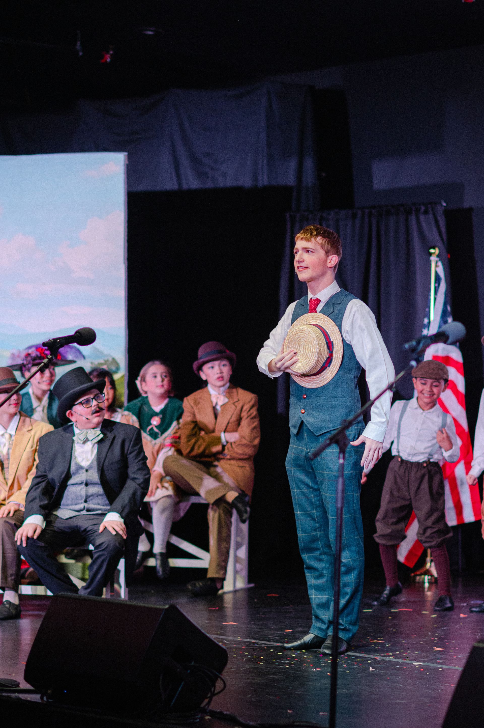 Man on stage holding a tambourine, with seated musicians and a red curtain backdrop from The Music Man