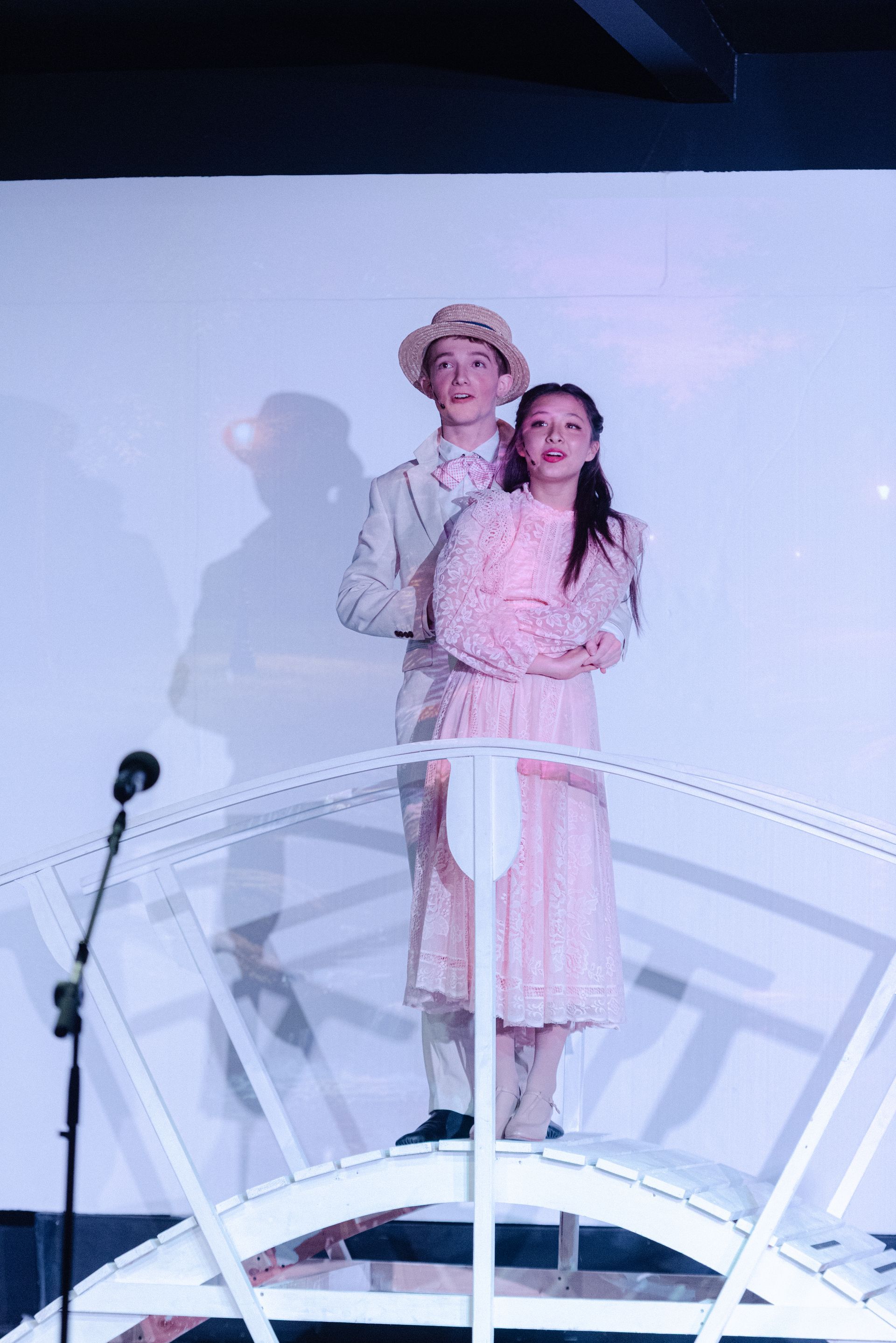 Two performers on a stage, standing close together in pastel outfits behind a microphone from The Music Man