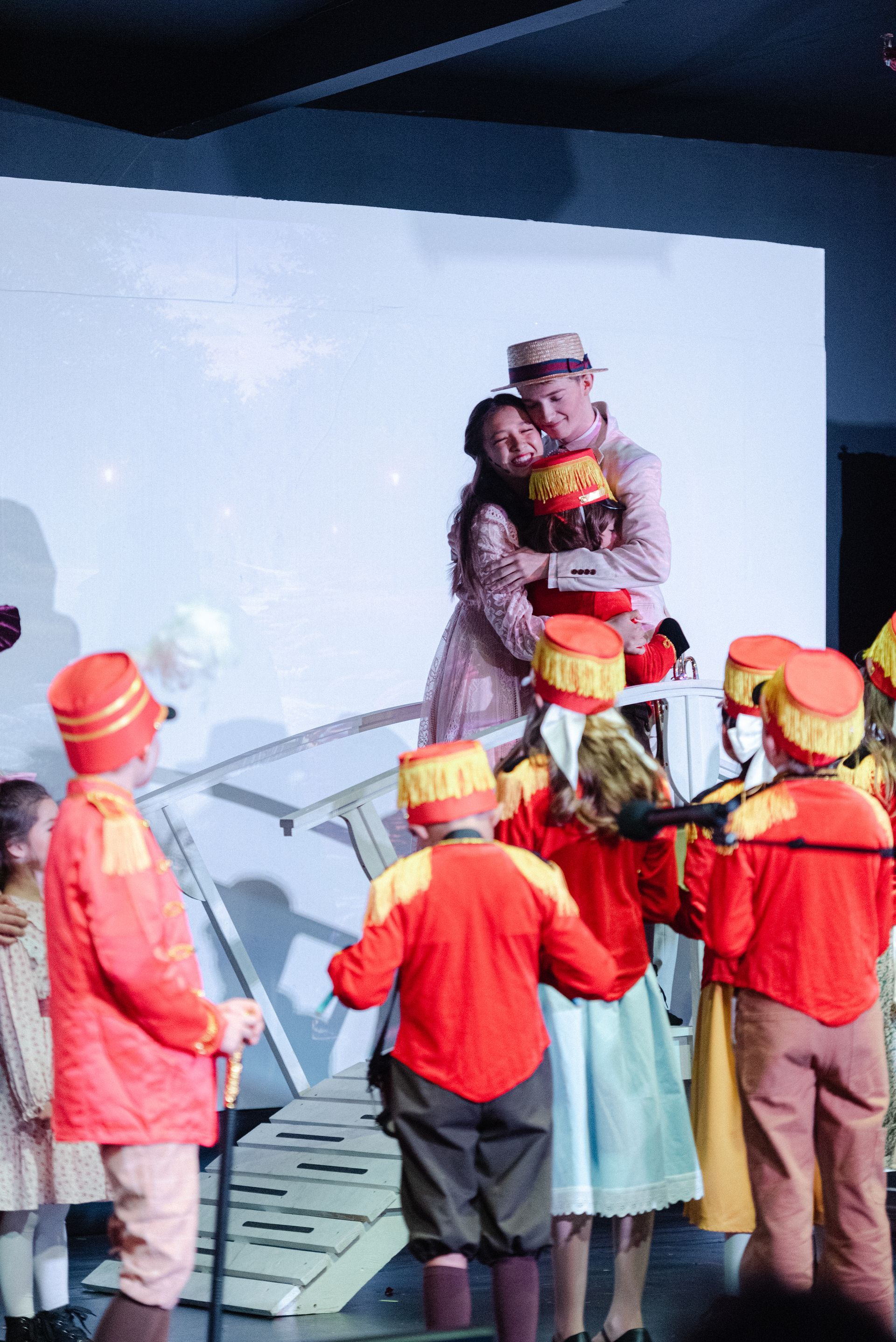 Children in red costumes gather around a person holding a wrapped gift onstage with a white backdrop from The Music Man