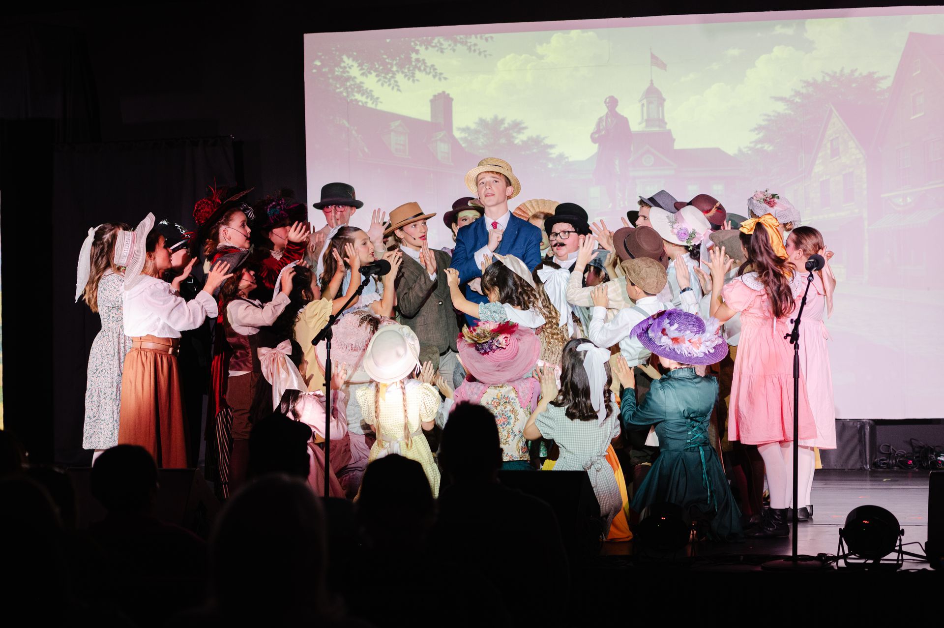 Costumed performers on a stage with microphones, flowers, and a projected backdrop, celebrating together from The Music Man
