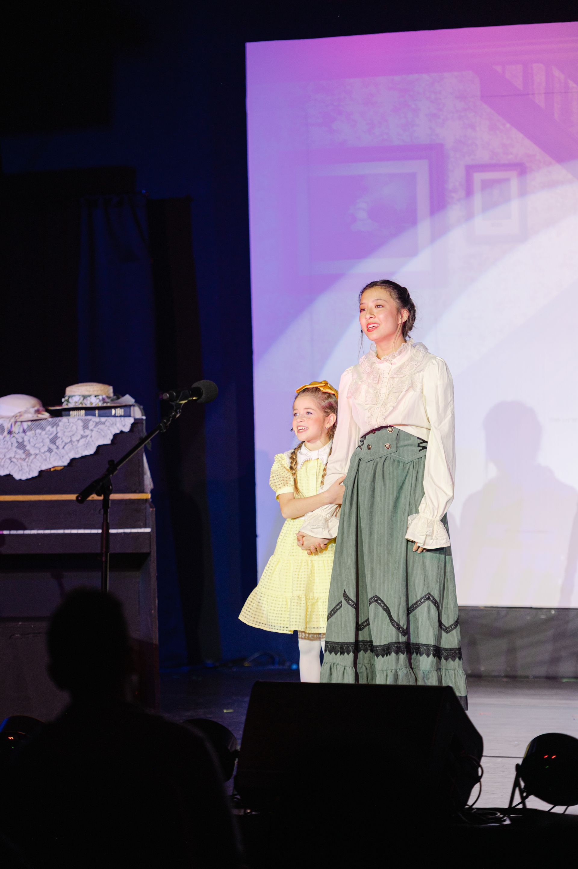 Performer and child on stage in formal costumes beside a piano, with a purple-lit backdrop from The Music Man