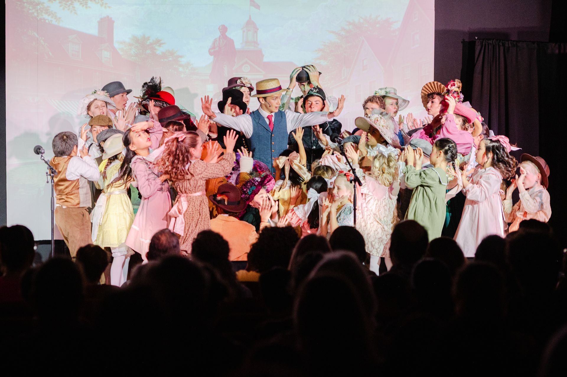 Large group of performers in colorful costumes on stage under pink lighting, seen from the audience from The Music Man