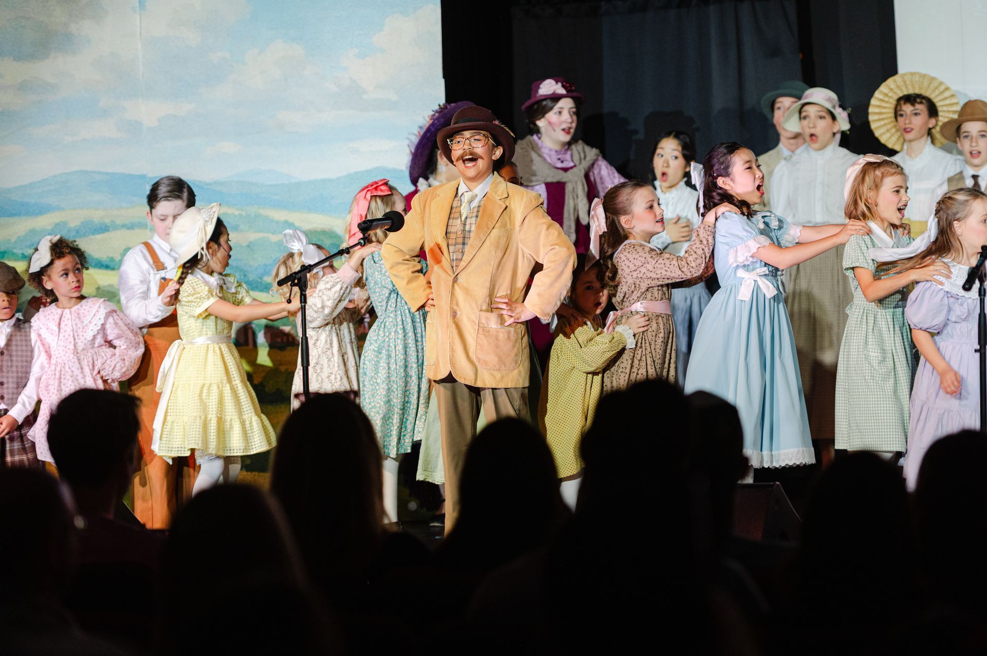 Theater cast performing on stage in colorful period costumes, with audience silhouettes in front from The Music Man