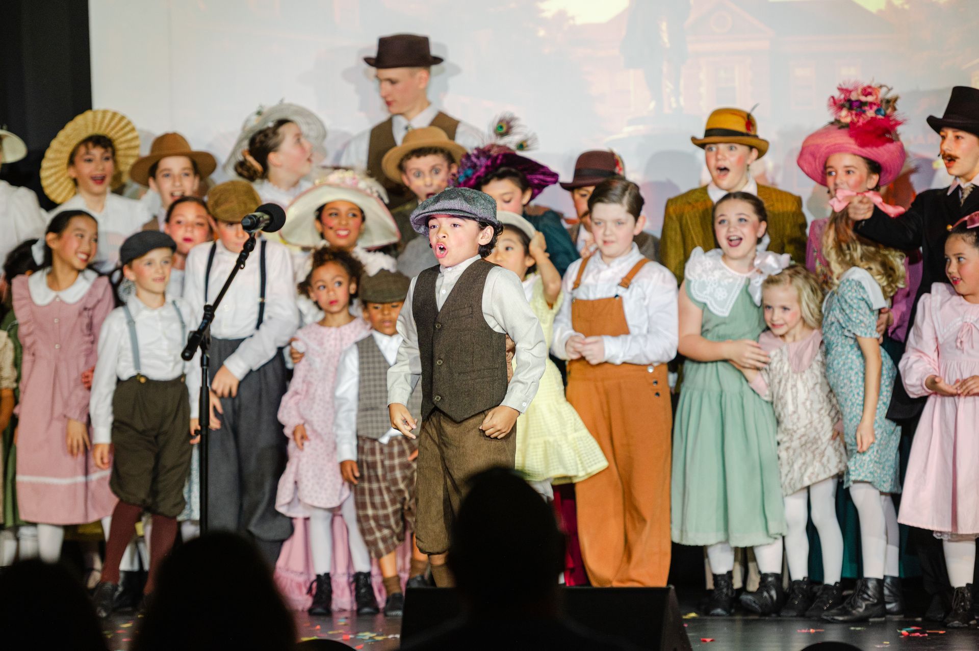 Children in vintage costumes stand on a stage during a performance, with a man in a top hat behind them from The Music Man