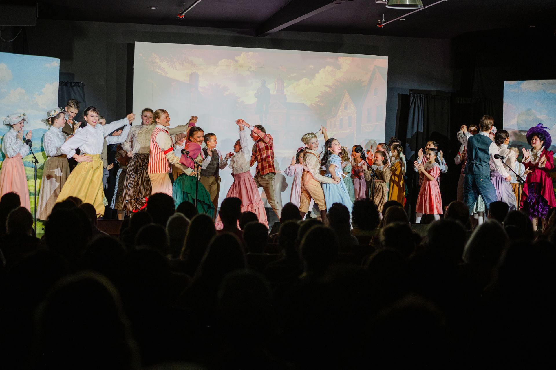 Large group of performers in colorful costumes on a stage, with audience silhouettes in front from The Music Man