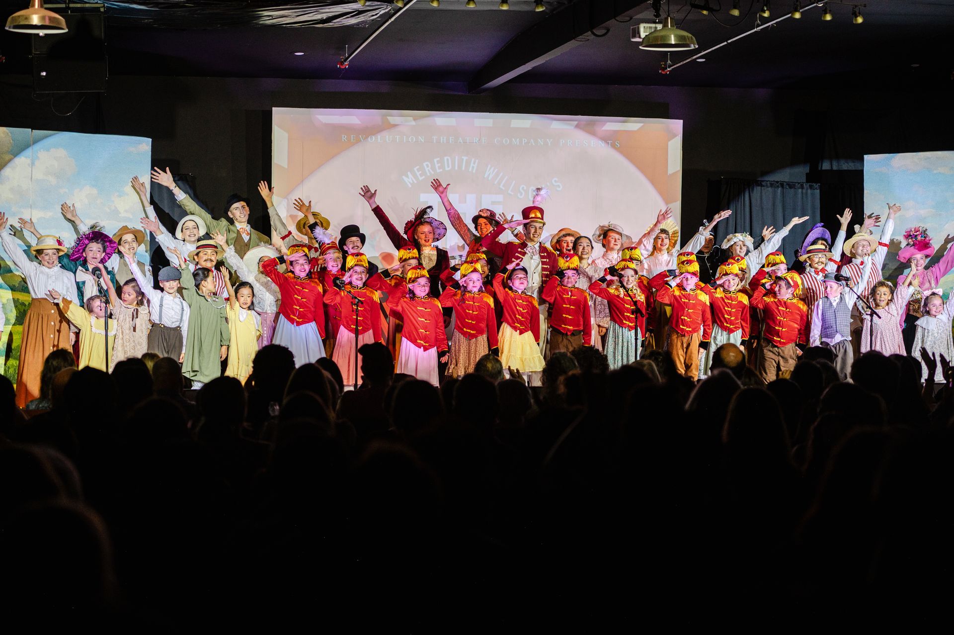 Large group of performers in colorful costumes onstage with raised arms under bright lights from The Music Man