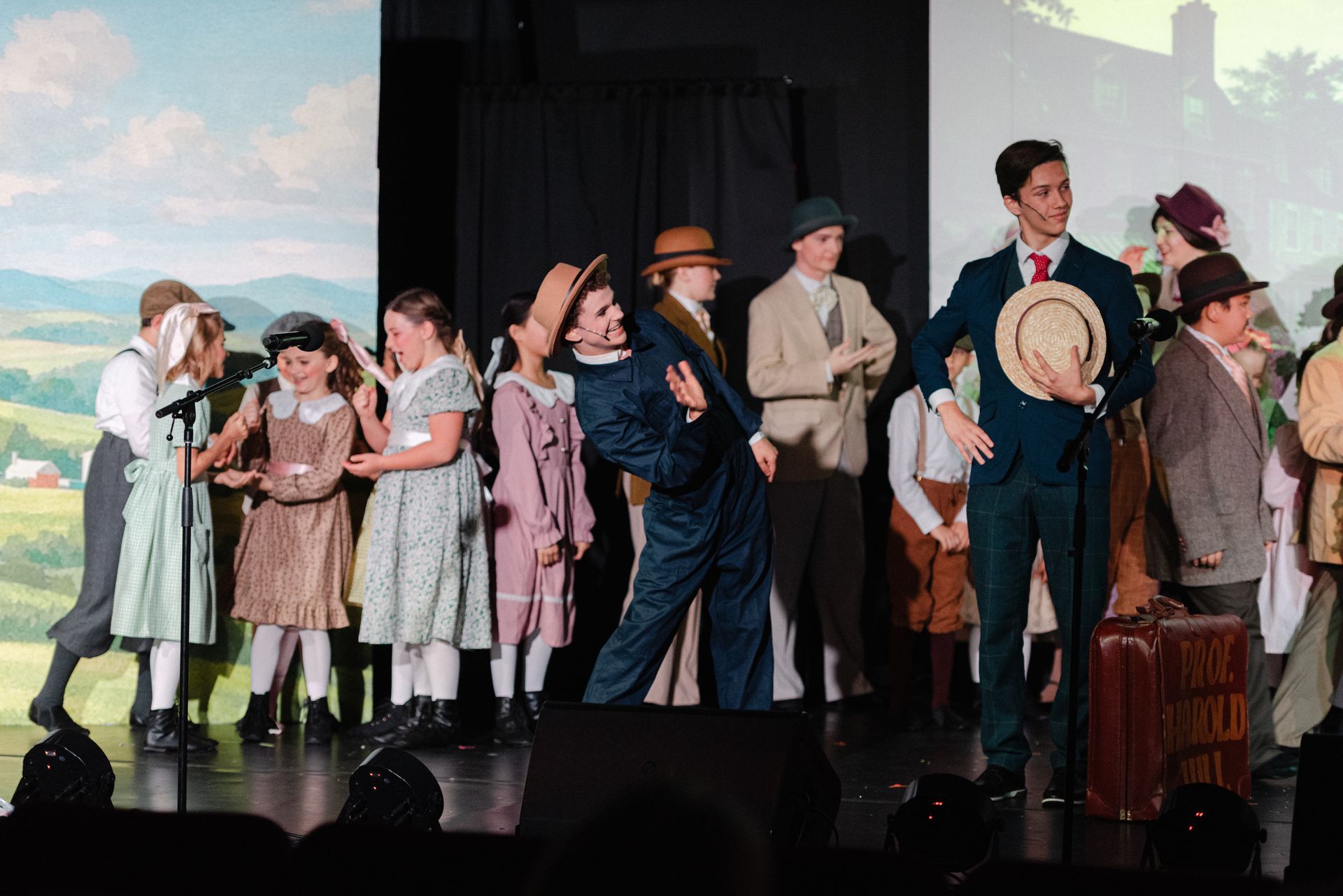 A group of people in period costumes stand on a stage in front of painted scenic backdrops from The Music Man
