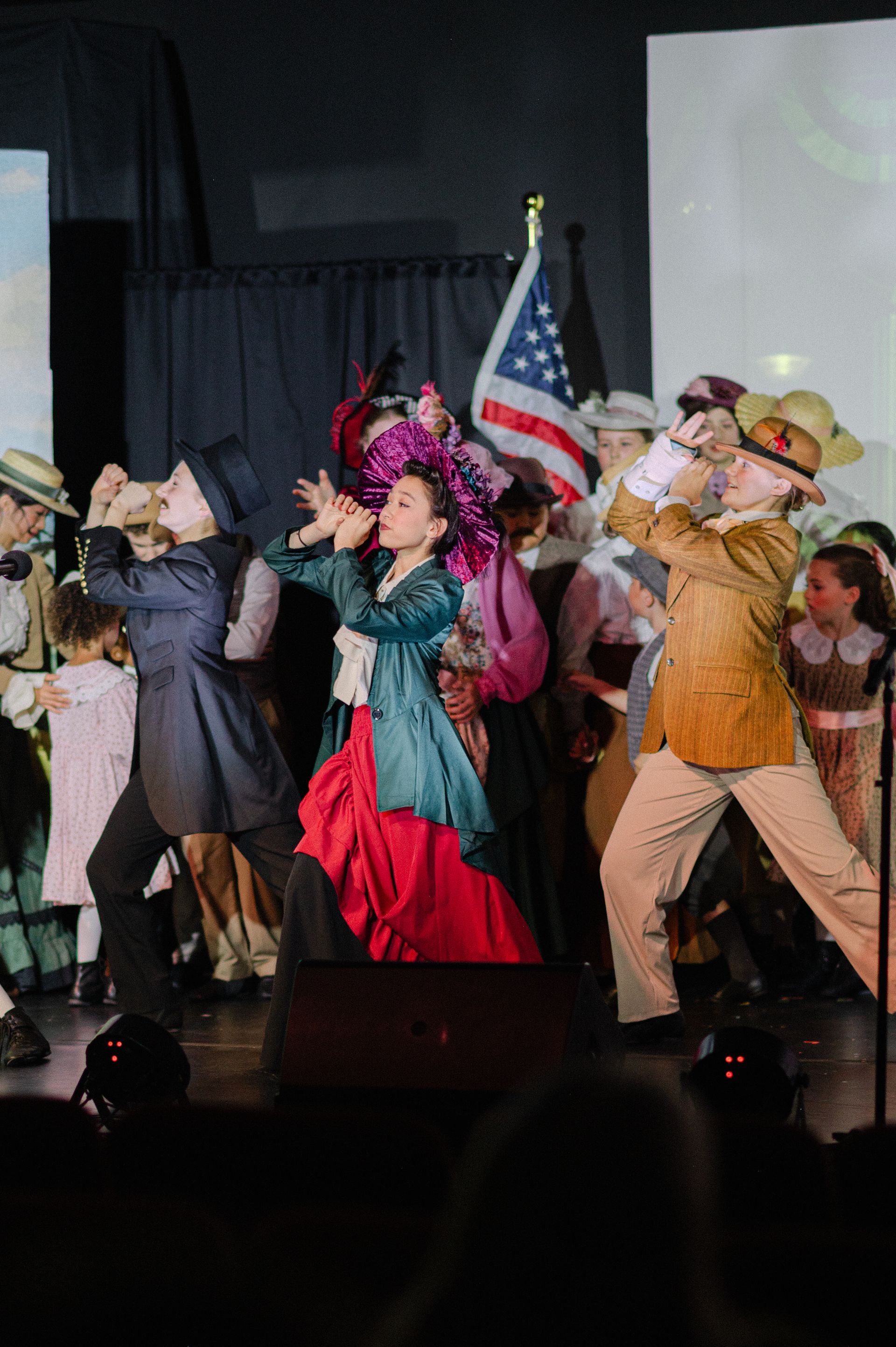 Performers dancing on stage with an American flag backdrop, wearing colorful costumes from The Music Man