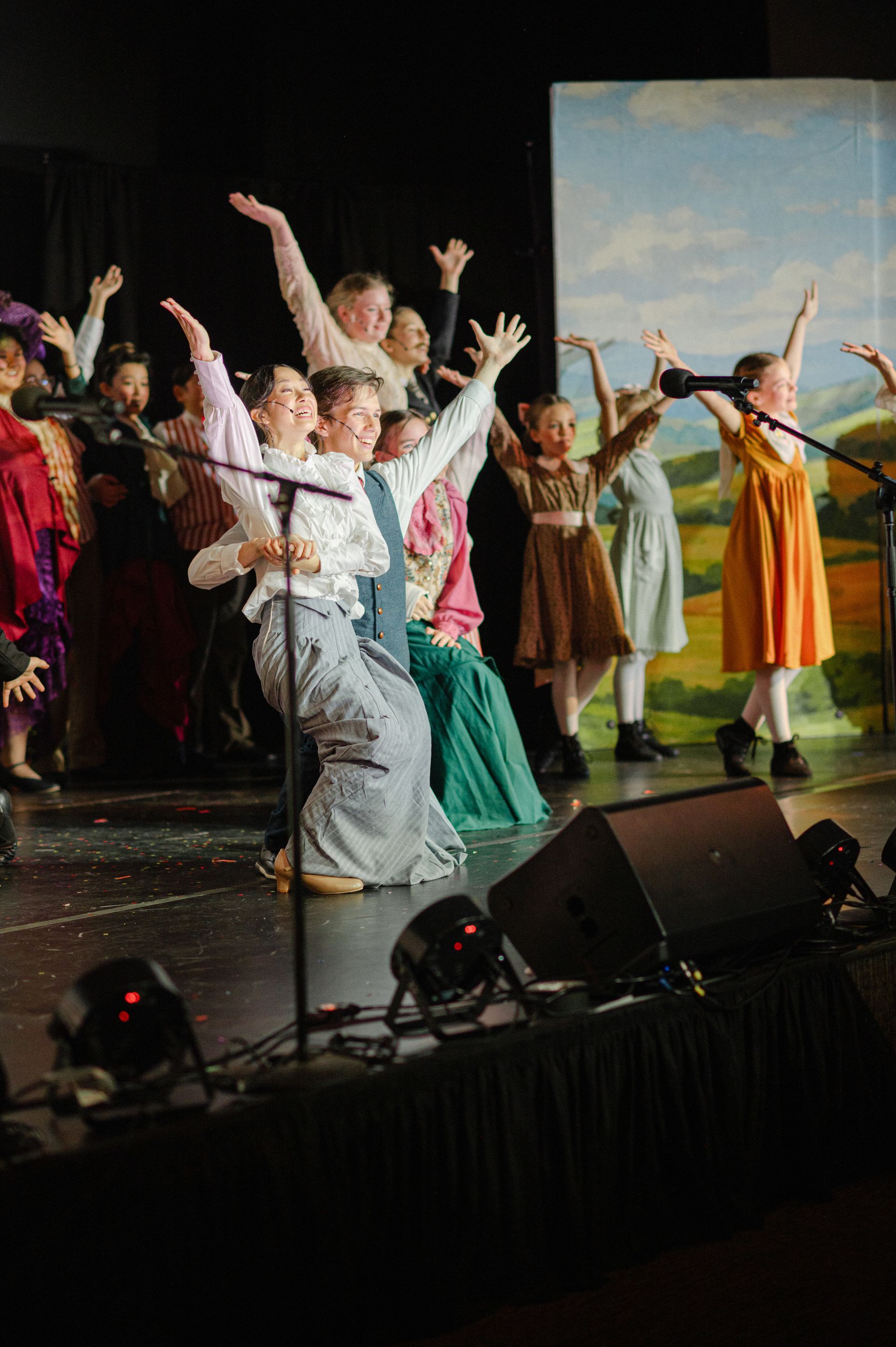 Performers in colorful costumes dance on a stage with raised arms and a painted backdrop from The Music Man