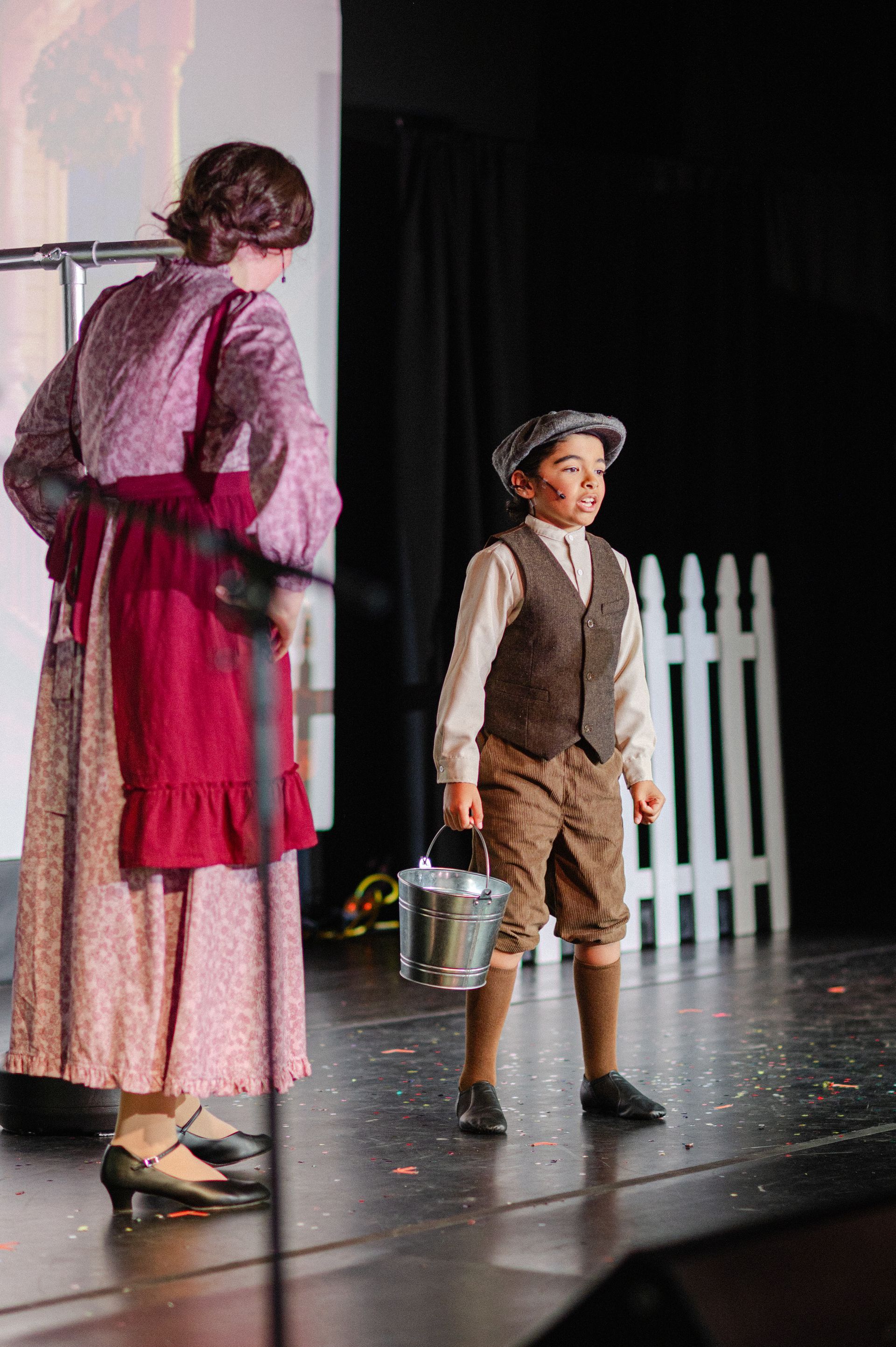 Child in a cap and vest stands onstage beside an adult in a pink dress; from The Music Man