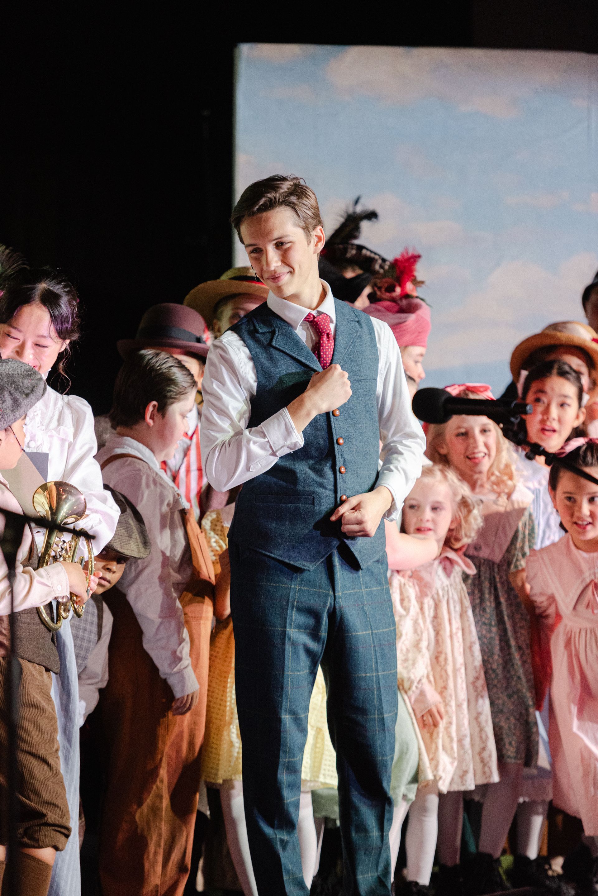 Man in blue suit stands center stage among children in costumes during a theatrical performance from The Music Man