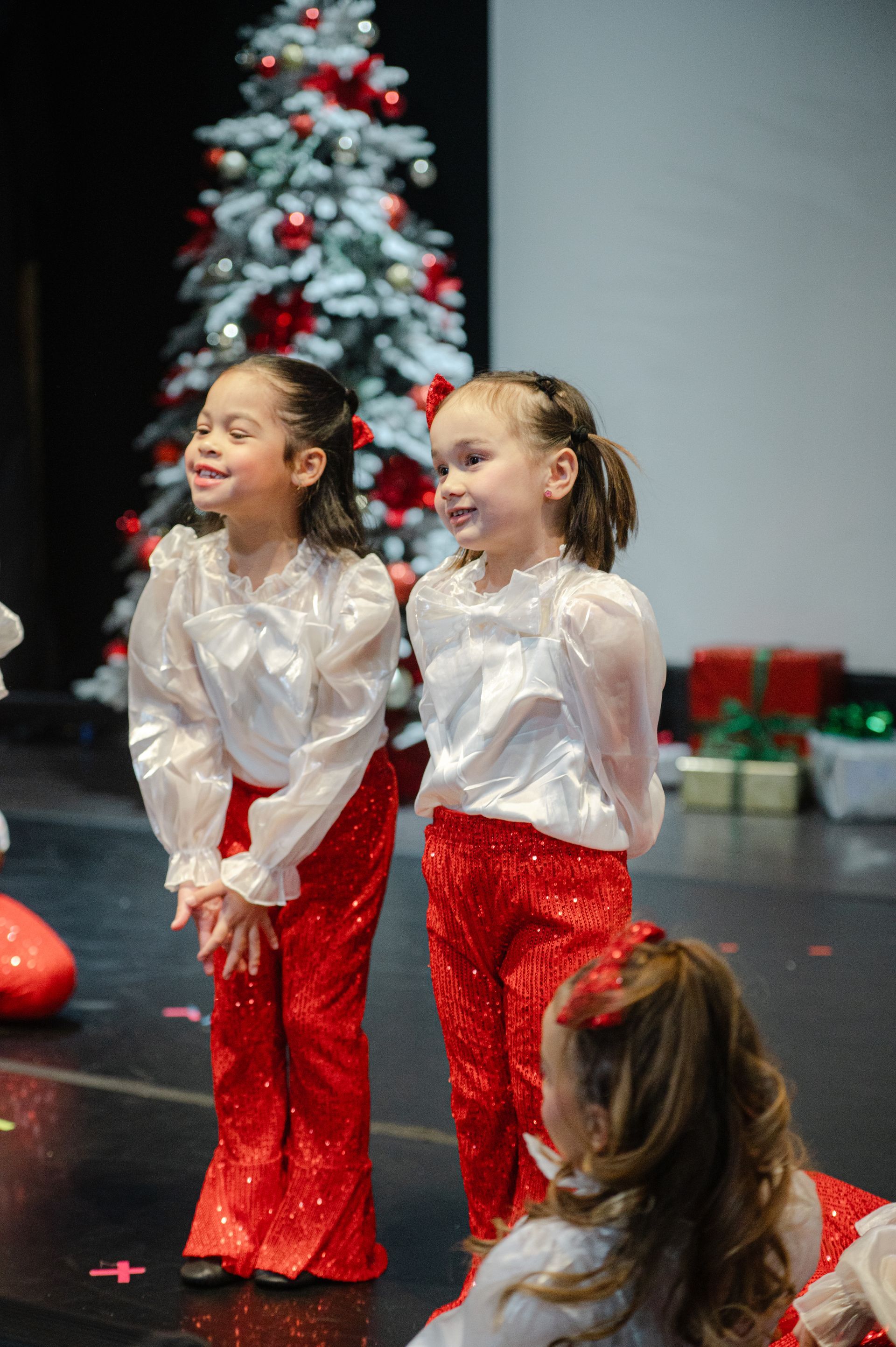 Two children in white tops and red pants perform onstage near a decorated Christmas tree.