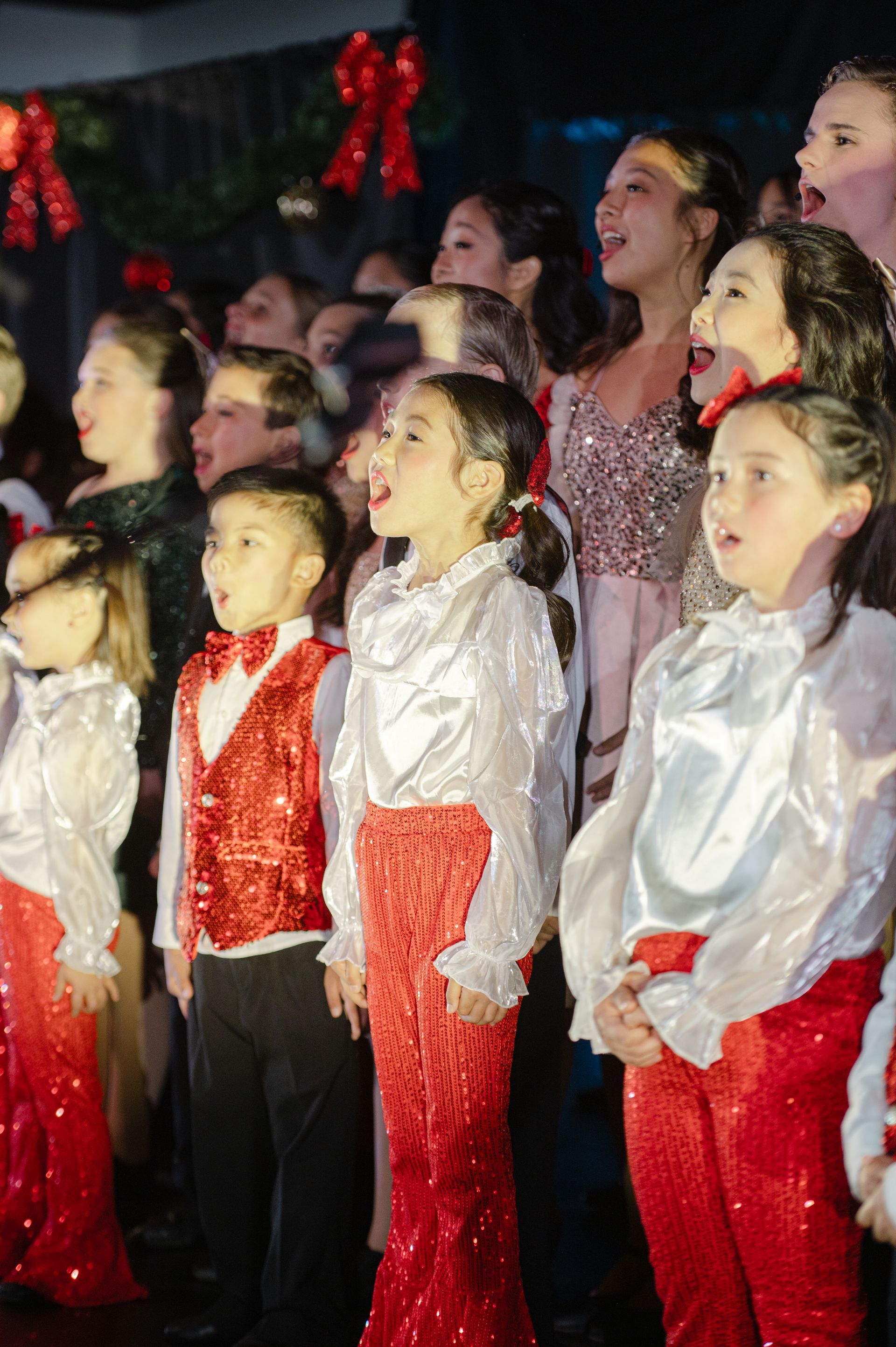 Children and adults singing on stage in festive red and white outfits at a holiday performance