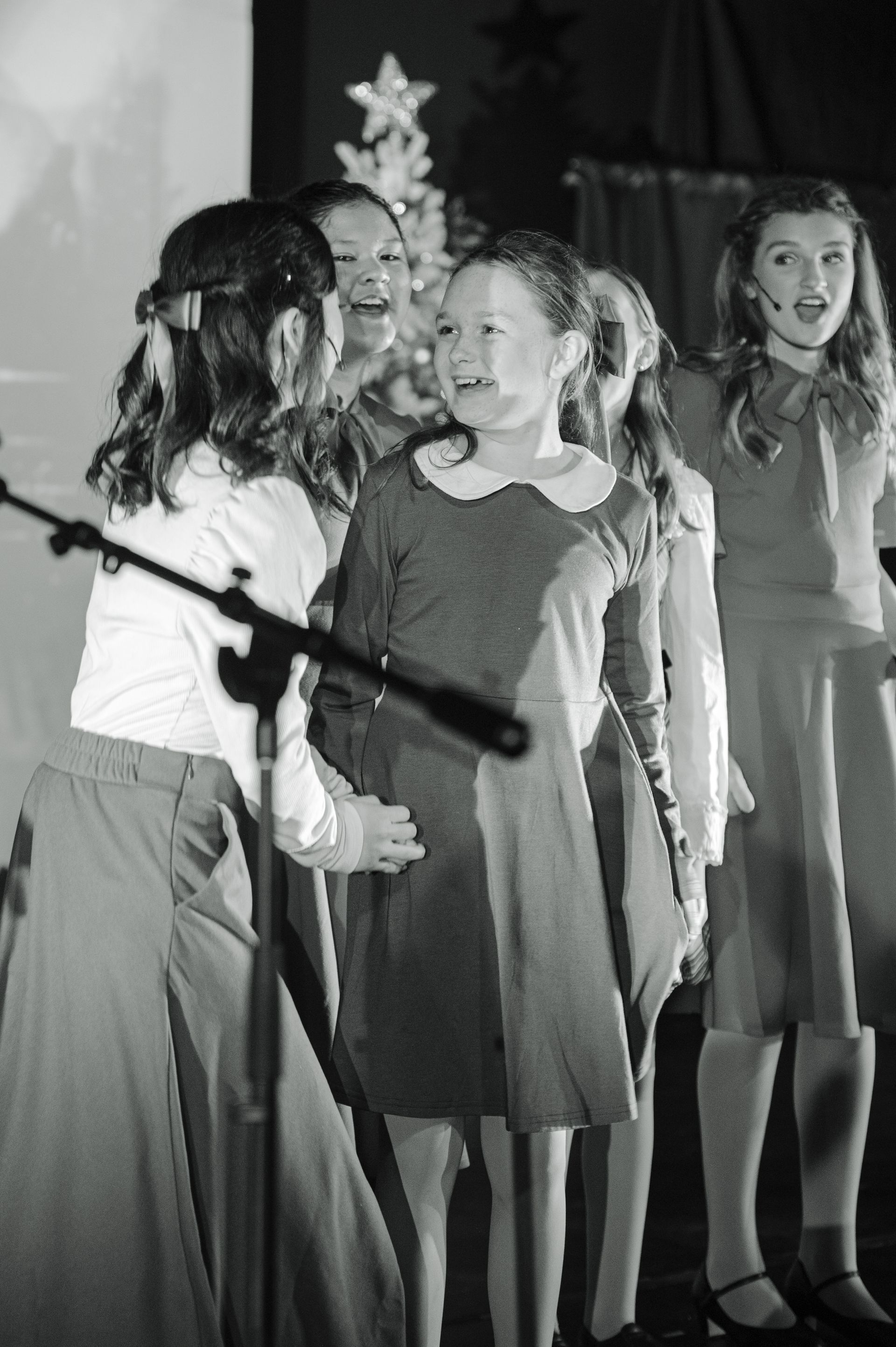 Children performing on stage, one singing into a microphone while others stand behind in dresses and sweaters