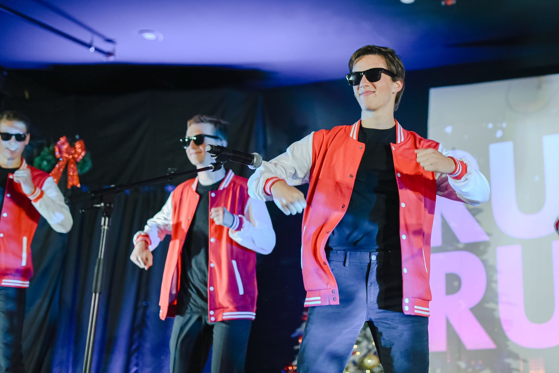 Three dancers in red jackets perform onstage under blue-purple lights, wearing sunglasses.