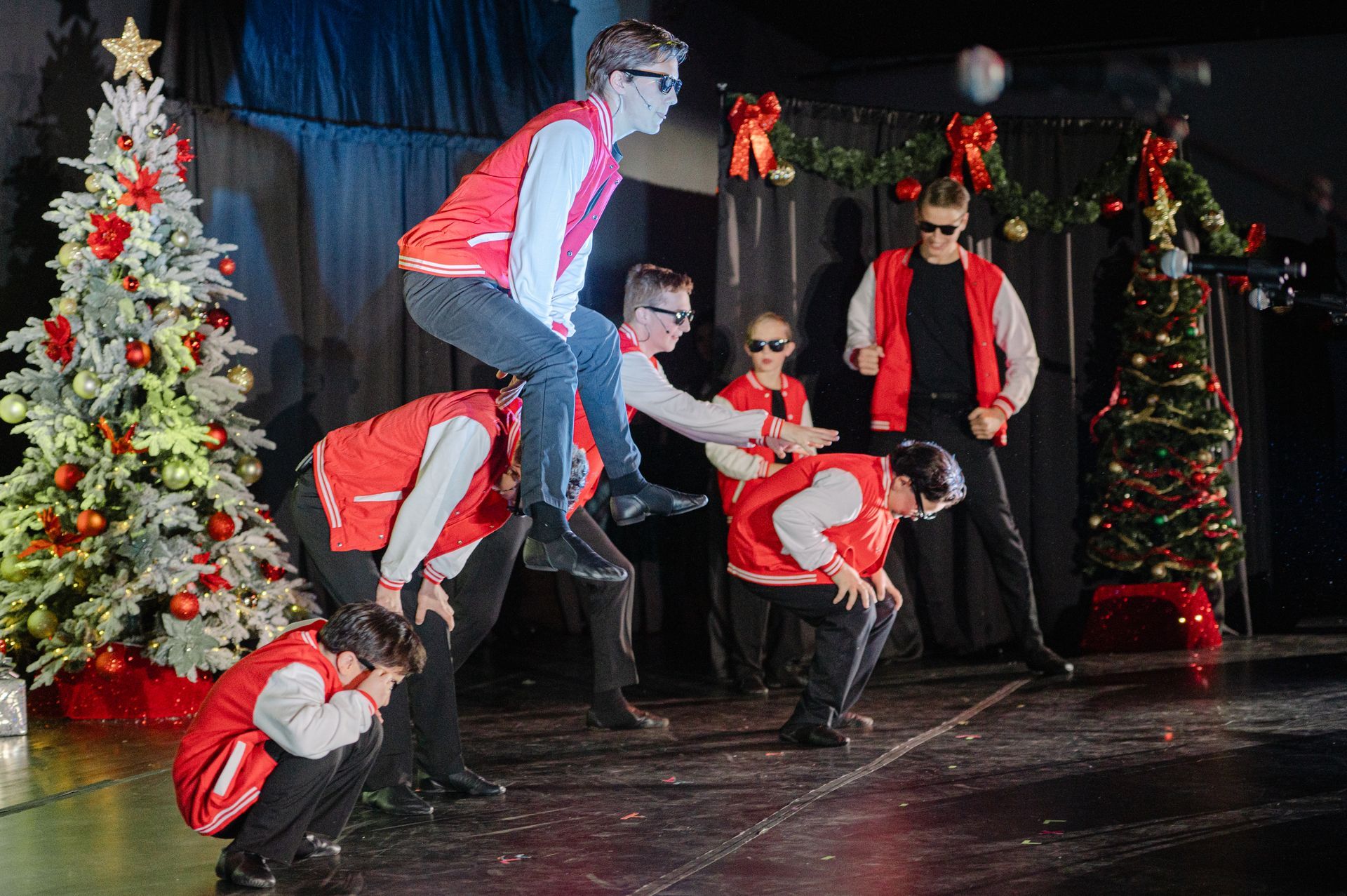 Performers in red vests pose in a festive stage scene with decorated Christmas trees and garlands