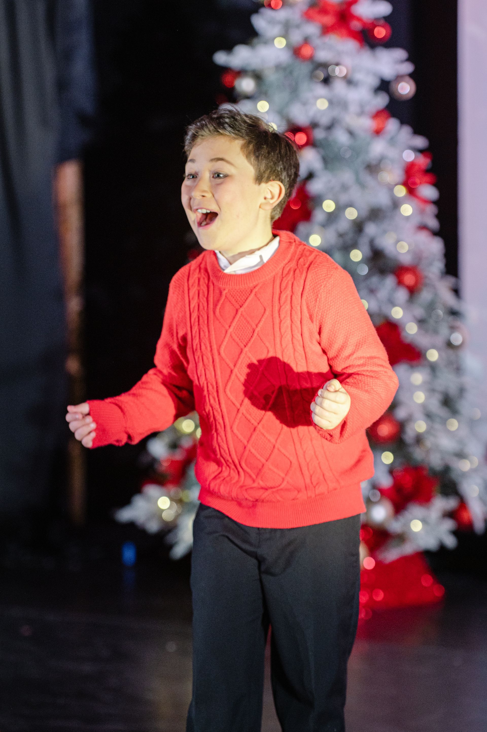 Person in a red sweater performing onstage in front of a lit Christmas tree