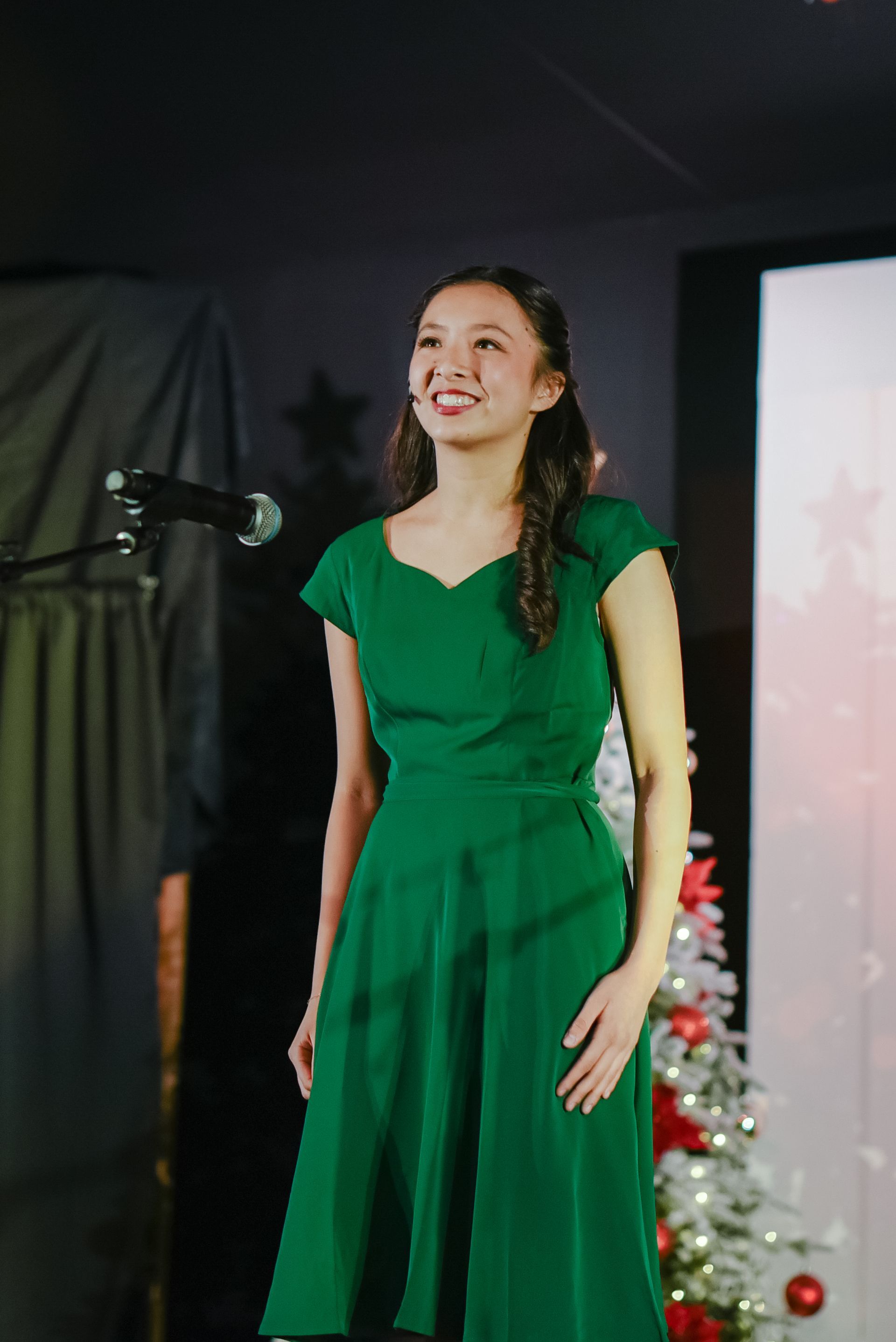 Woman in a green dress singing on stage beside a microphone, with a decorated Christmas tree in the background