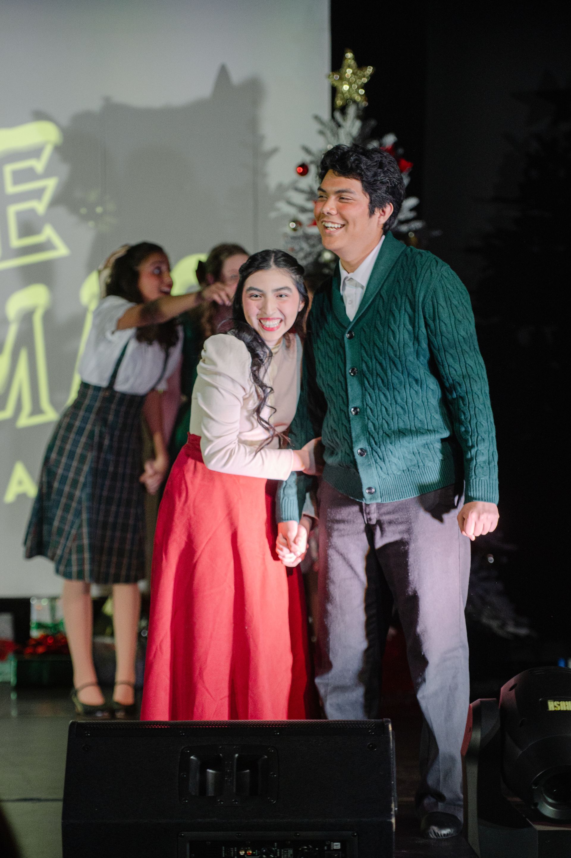 Two people smiling on stage, one in a red skirt and green sweater, with a star decoration behind them.