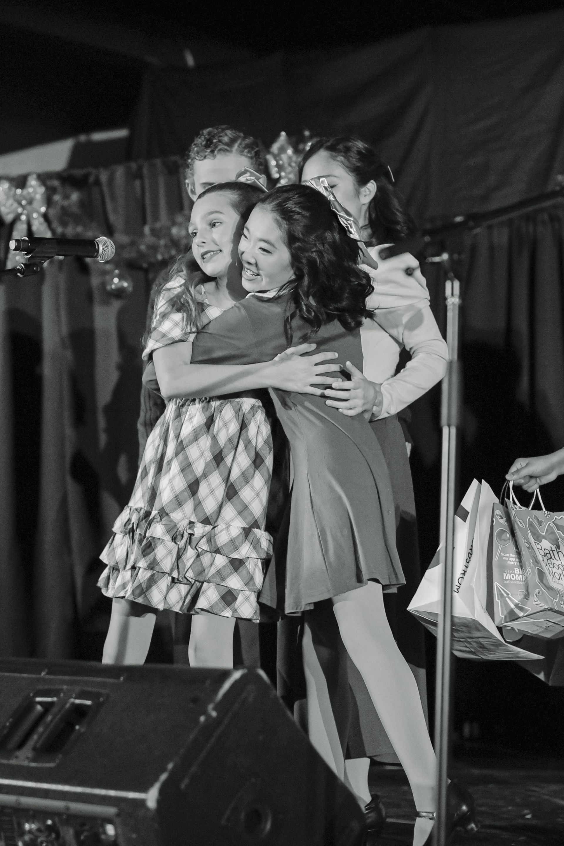 Four people hugging on a stage, smiling in a black-and-white photo.