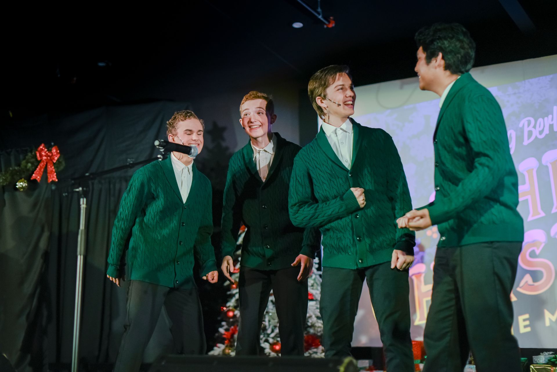 Four men in green blazers smiling onstage during a performance or presentation