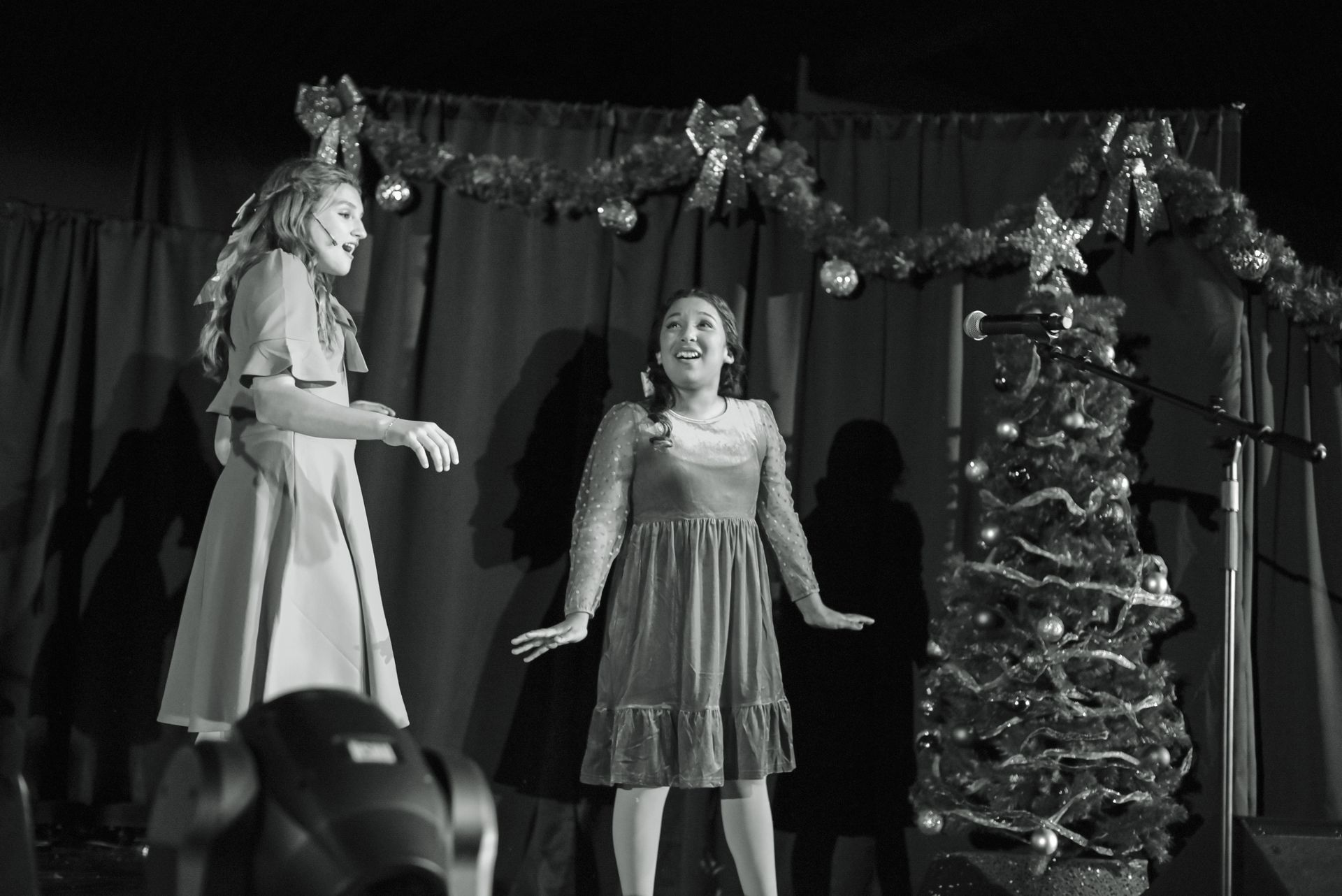 Two performers onstage beside a decorated Christmas tree and garlands, one singing into a microphone.